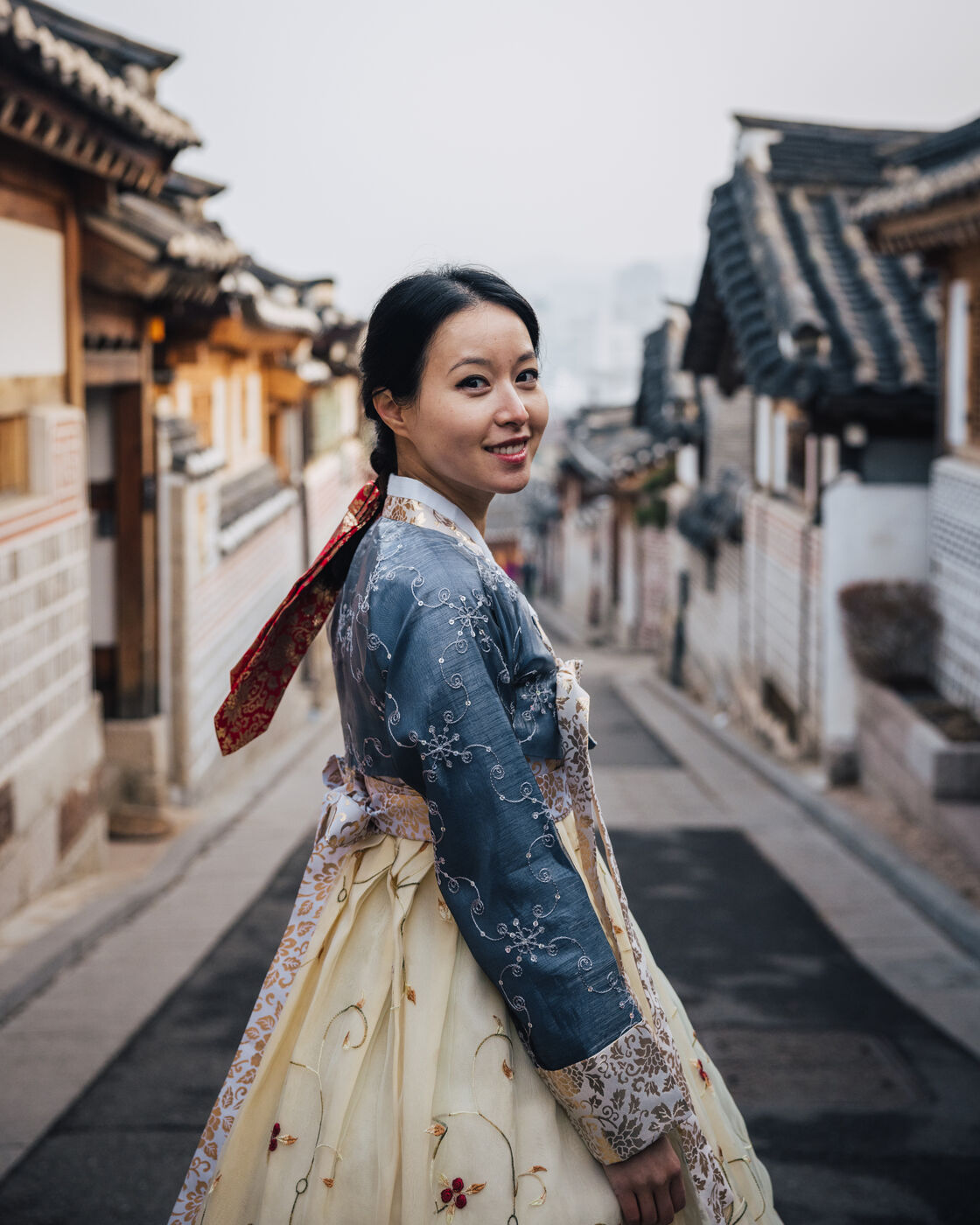 Grace Chen in a traditional Korean hanbok, looking back with a warm smile on a quiet street lined with traditional hanok houses in Seoul's Bukchon village