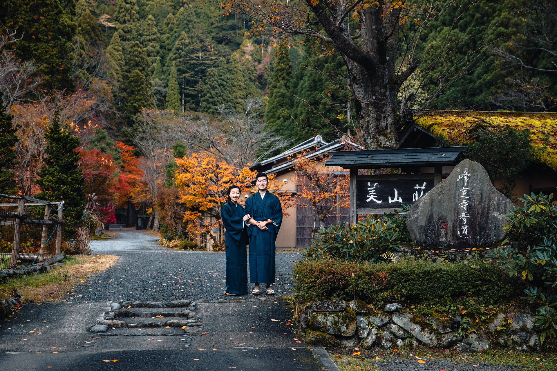 Jason Wang and Grace Chen in indigo kimonos at the entrance of Miyamasou ryokan, framed by autumn maples in the mountains north of Kyoto