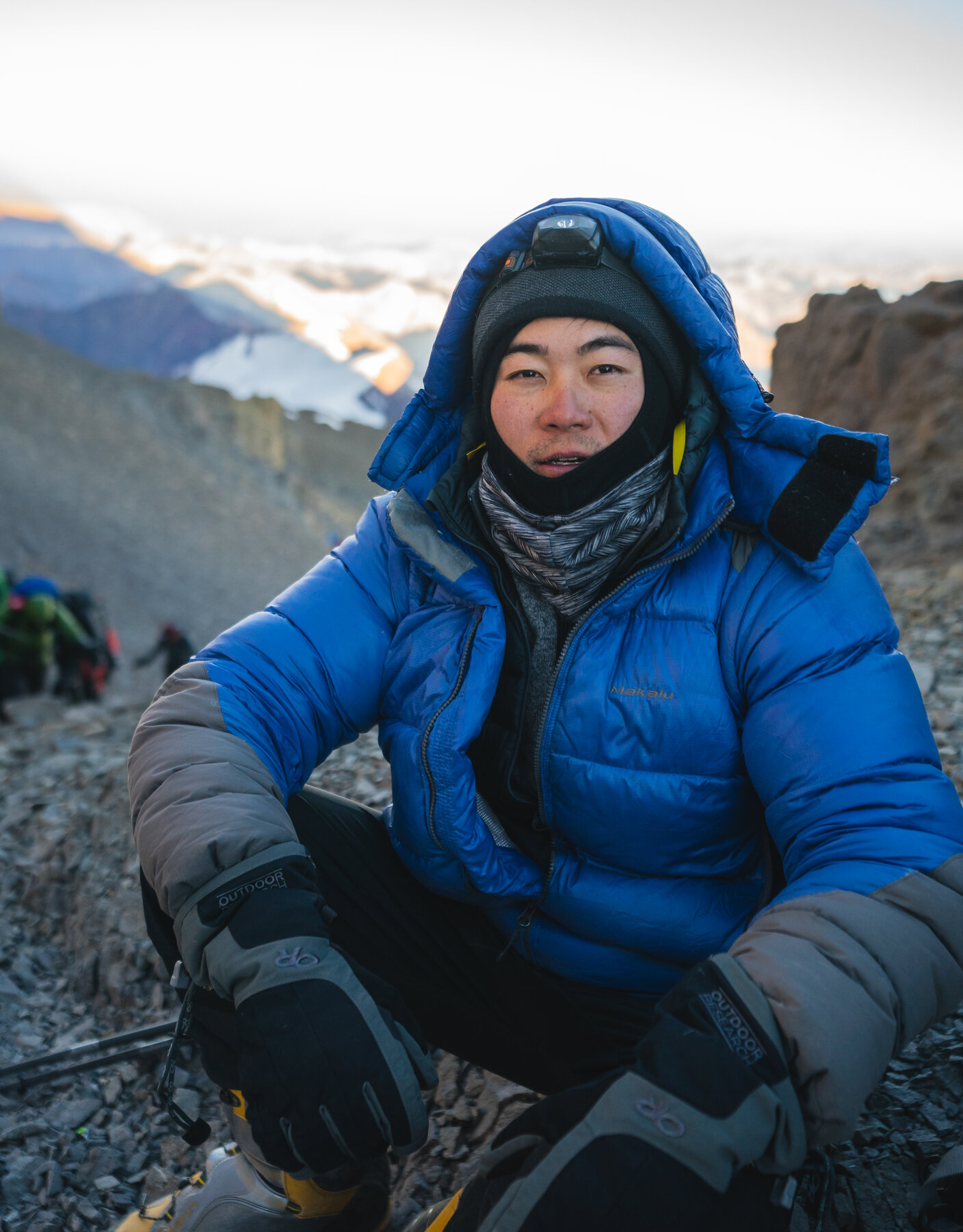 Grace sitting on rocks near the Aconcagua summit in a blue down jacket with mountains behind