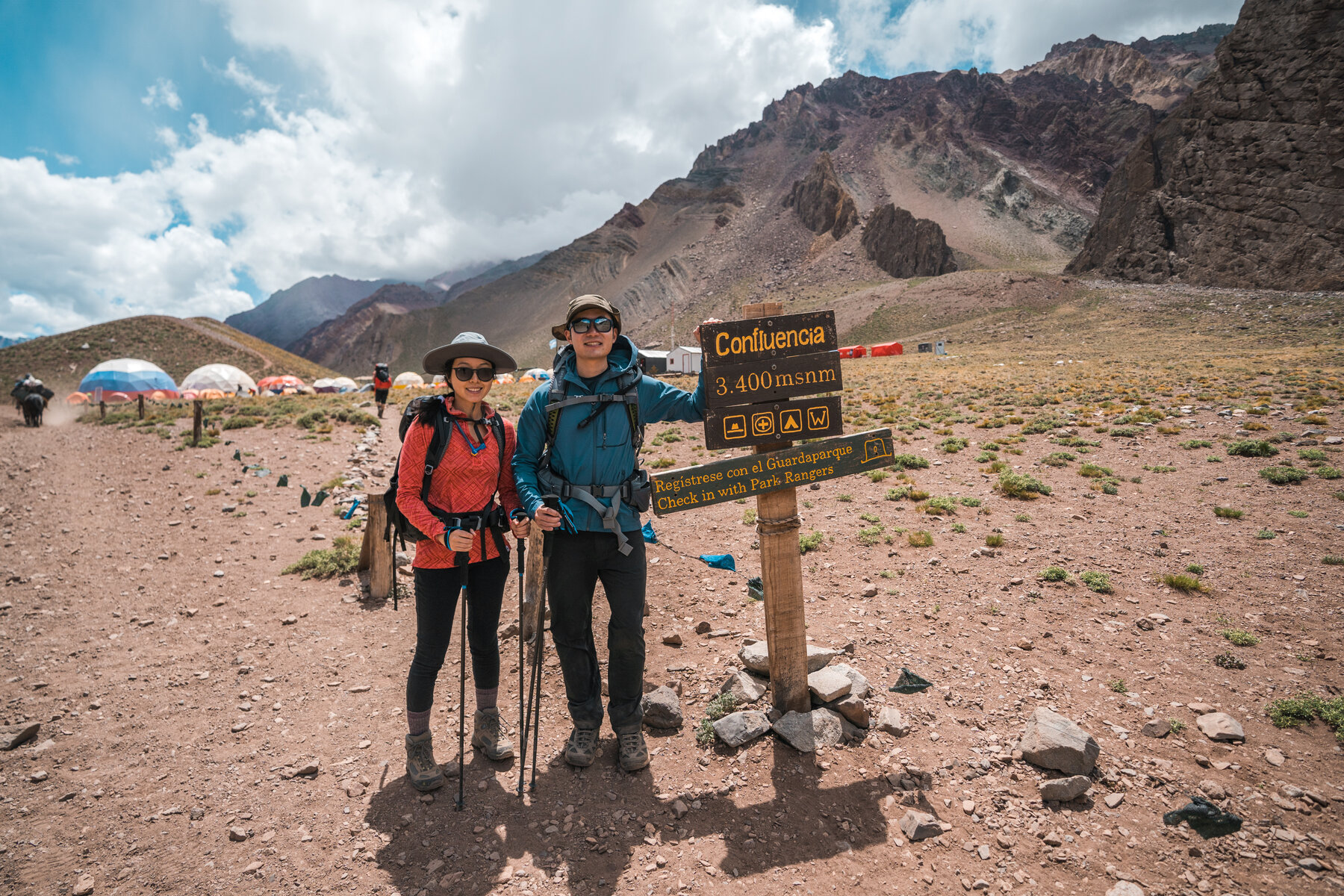 Jason and Grace at the Confluencia Camp waypoint sign with colorful tents behind