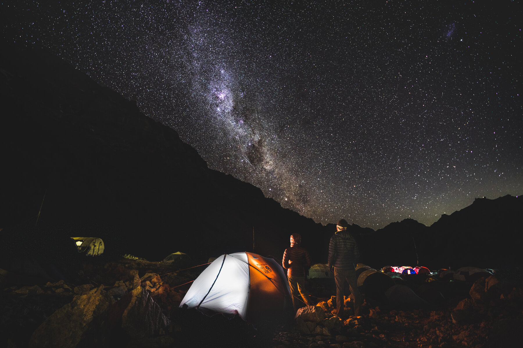 The Milky Way stretching over camp with a glowing tent and two climbers standing outside