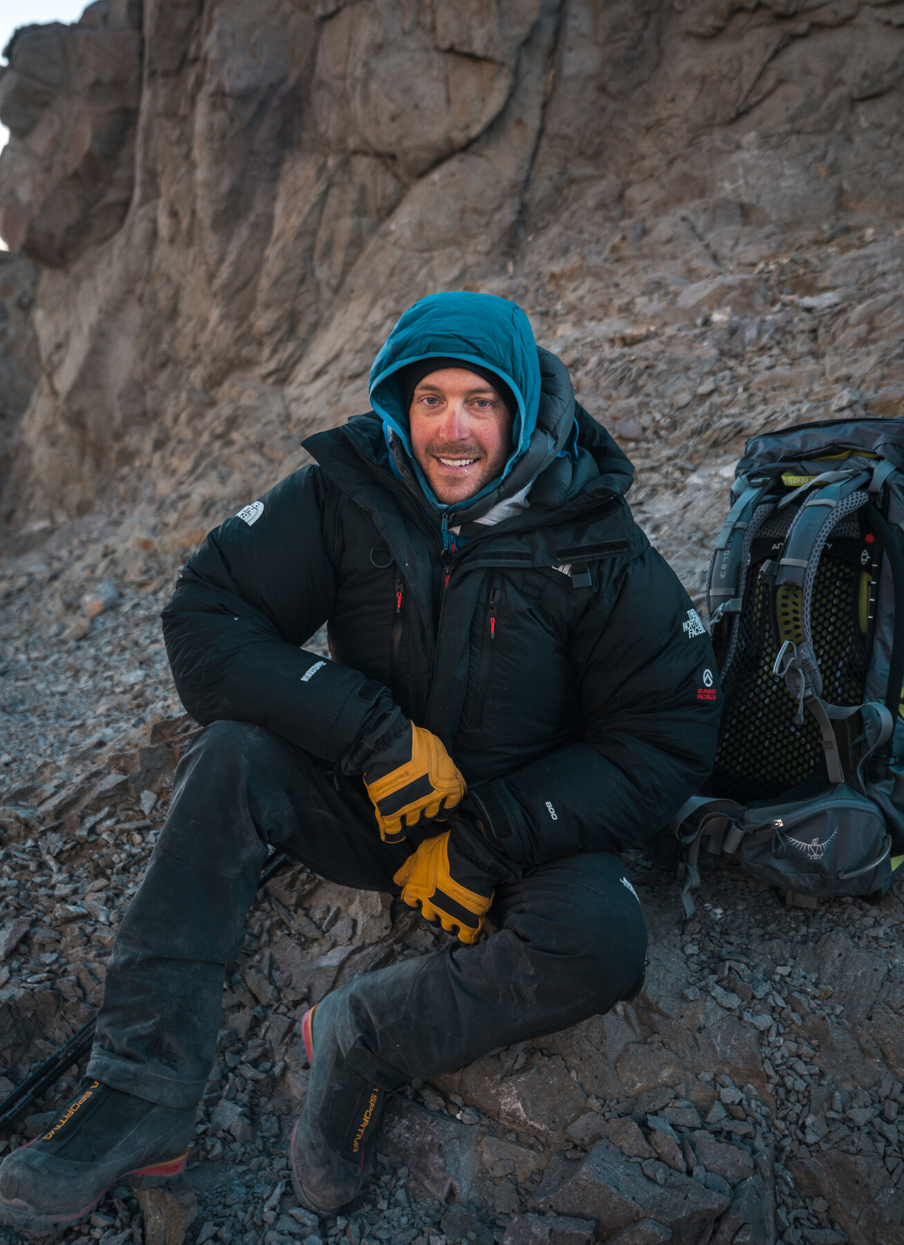Smiling team member in black North Face jacket and blue hood sitting on rocks near the summit