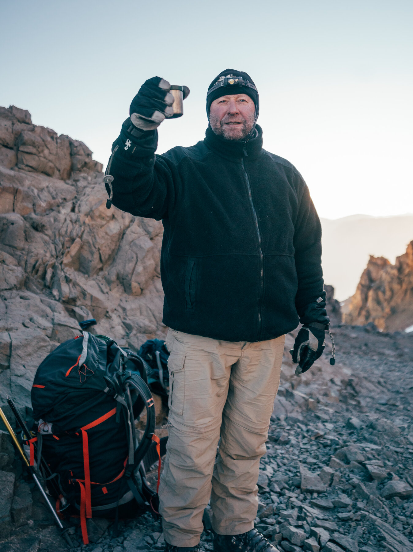 Bearded climber in a black jacket and beanie raising a gloved hand beside his pack on rocky ground high on the mountain