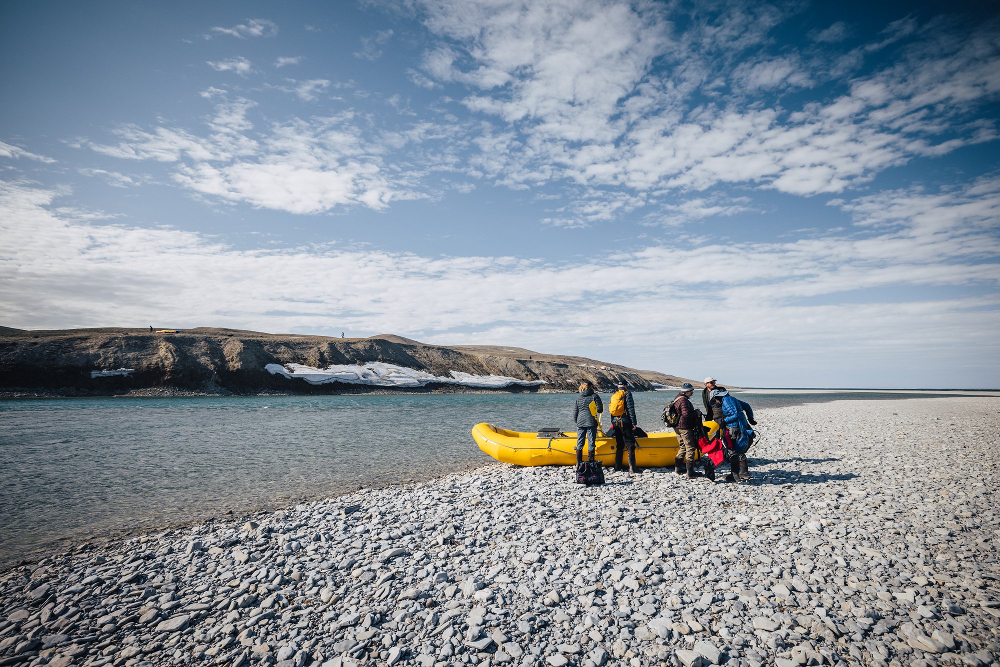 Guests with a yellow raft on a pebble beach, snow-streaked cliffs across the river