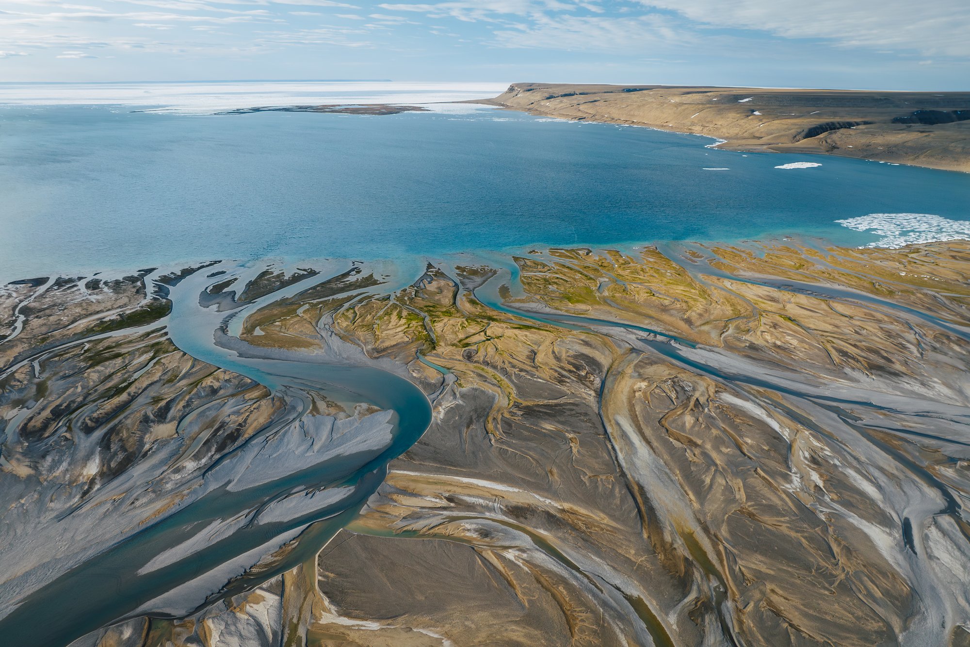 Aerial view of the Cunningham River delta's ochre and silver channels meeting a turquoise bay, sea ice on the horizon