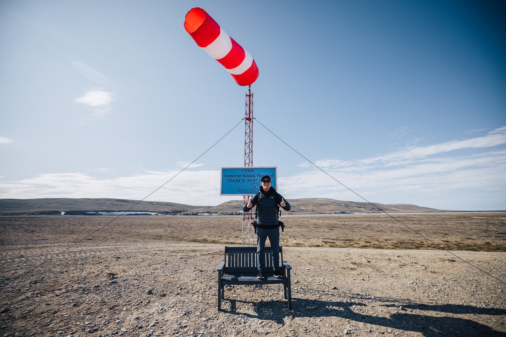 Jason on a bench beneath the red-and-white windsock at the CYRW airstrip sign, Somerset Island, 74 degrees 4 minutes north