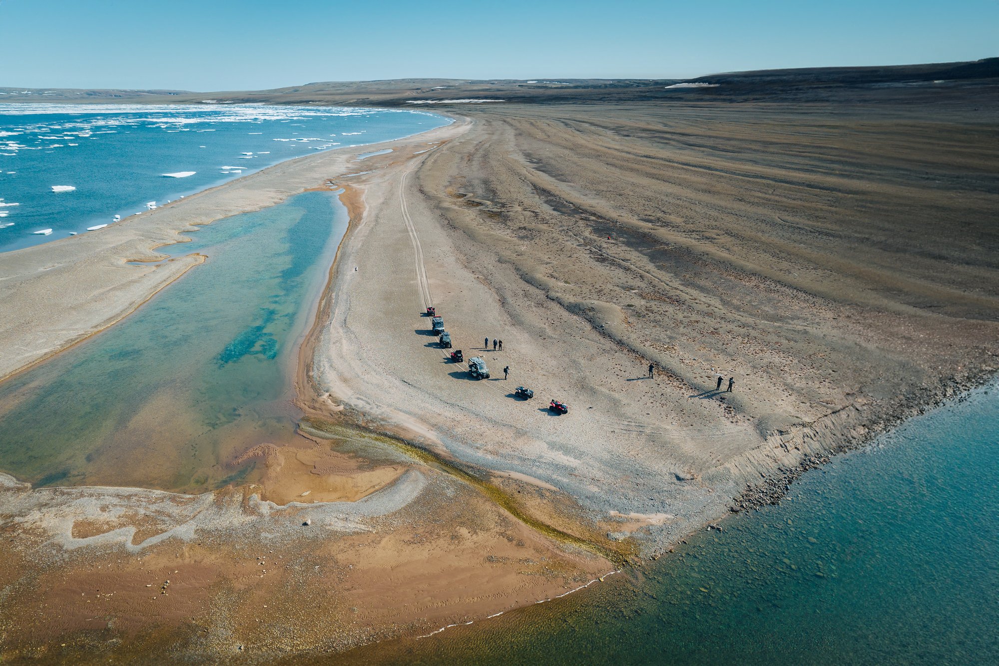 Aerial view of a long gravel spit with buggies parked, a turquoise lagoon on one side and the sea on the other