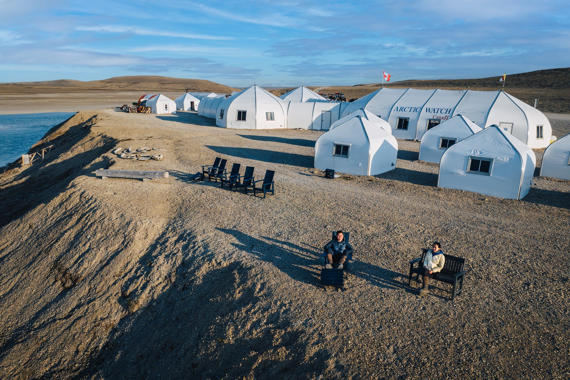 Jason and Grace in chairs outside Arctic Watch's white tents in low evening sun, whale bones on the gravel