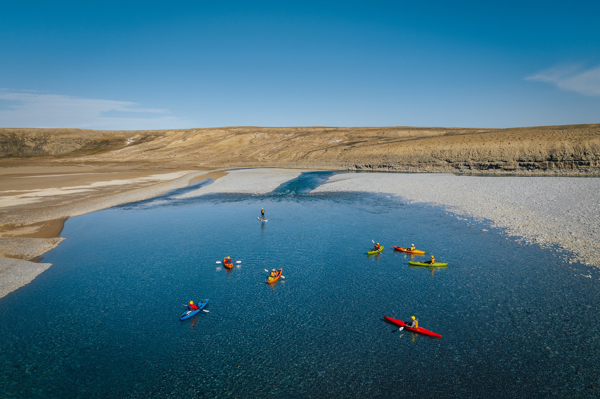A flotilla of kayaks and a stand-up paddleboarder on a wide blue stretch of the river