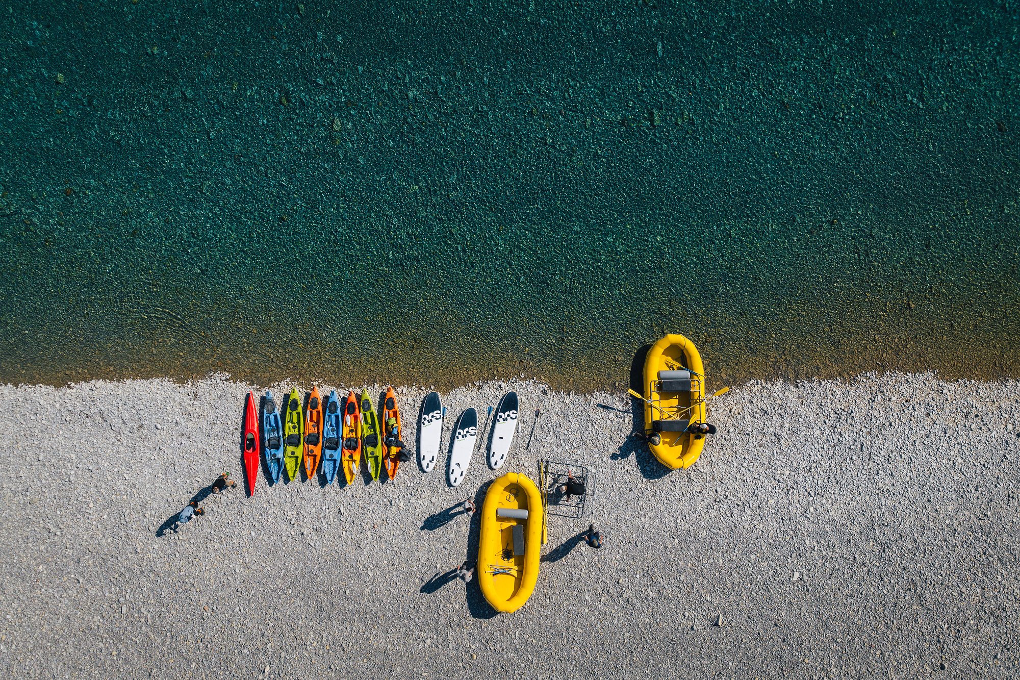 Top-down view of a row of kayaks, paddleboards and two yellow rafts lined up on a gravel bar beside green water