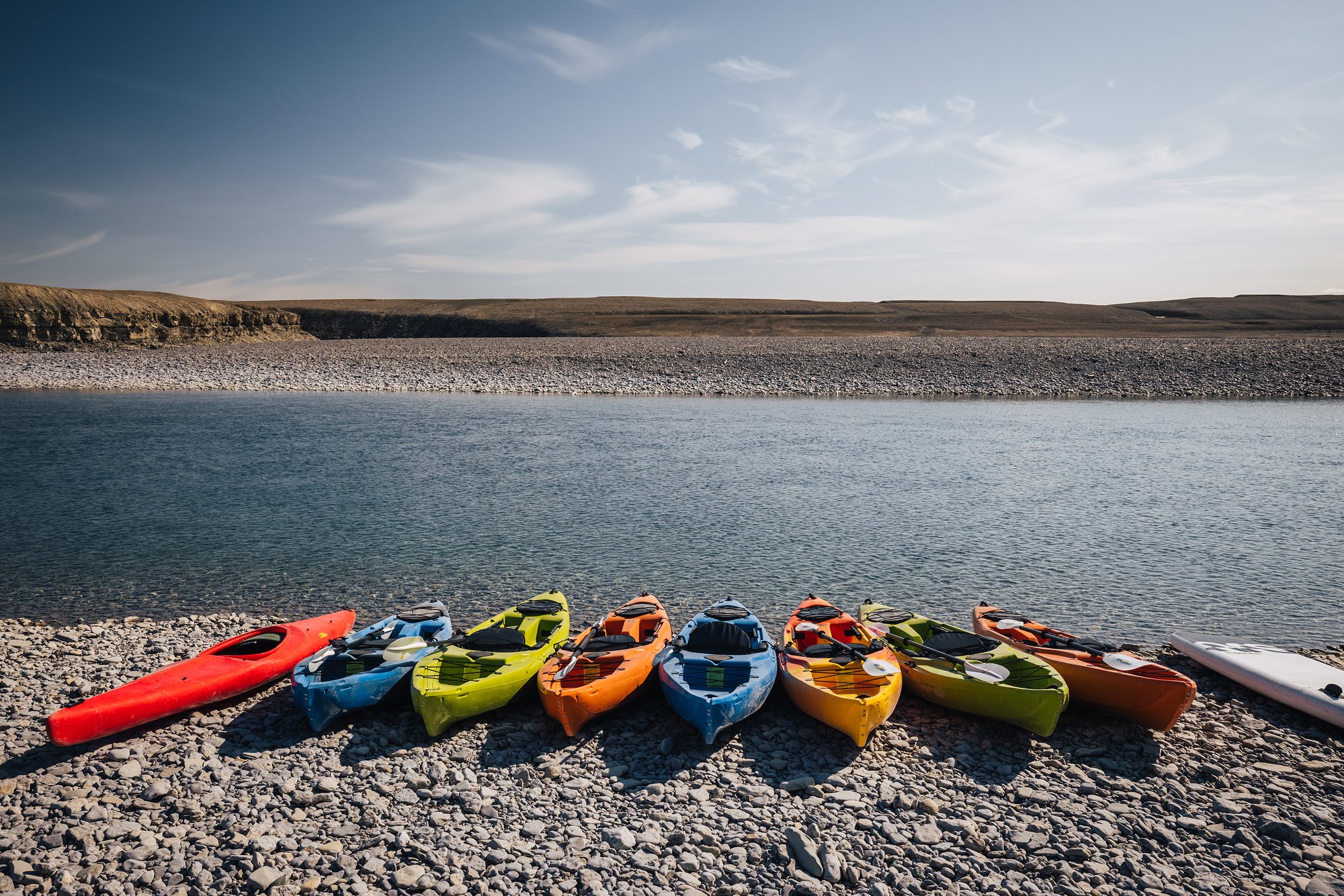 A row of colorful kayaks lined up on the pebble shore of the river