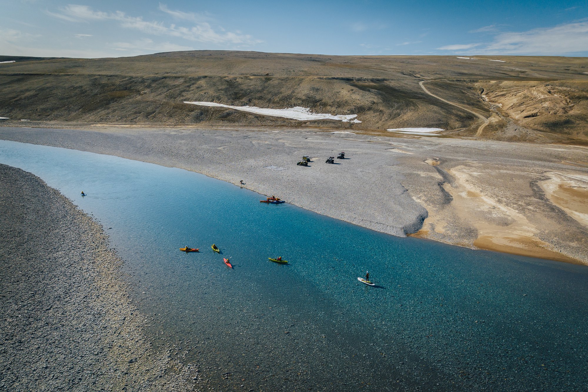 Aerial view of kayakers and rafts on a blue channel, ATVs parked on the gravel bar