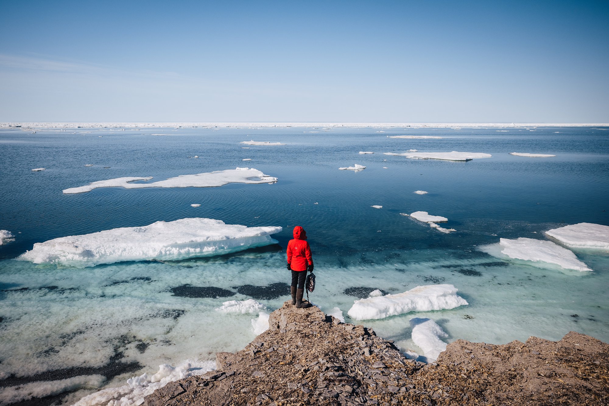 A guest in a red jacket on a rocky point looking out over ice floes on turquoise water