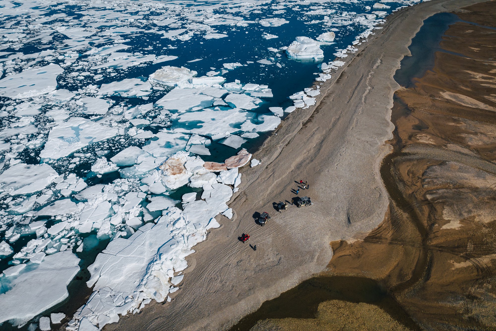 Aerial view of a group walking a gravel spit between a sea of broken ice floes and brown shallows