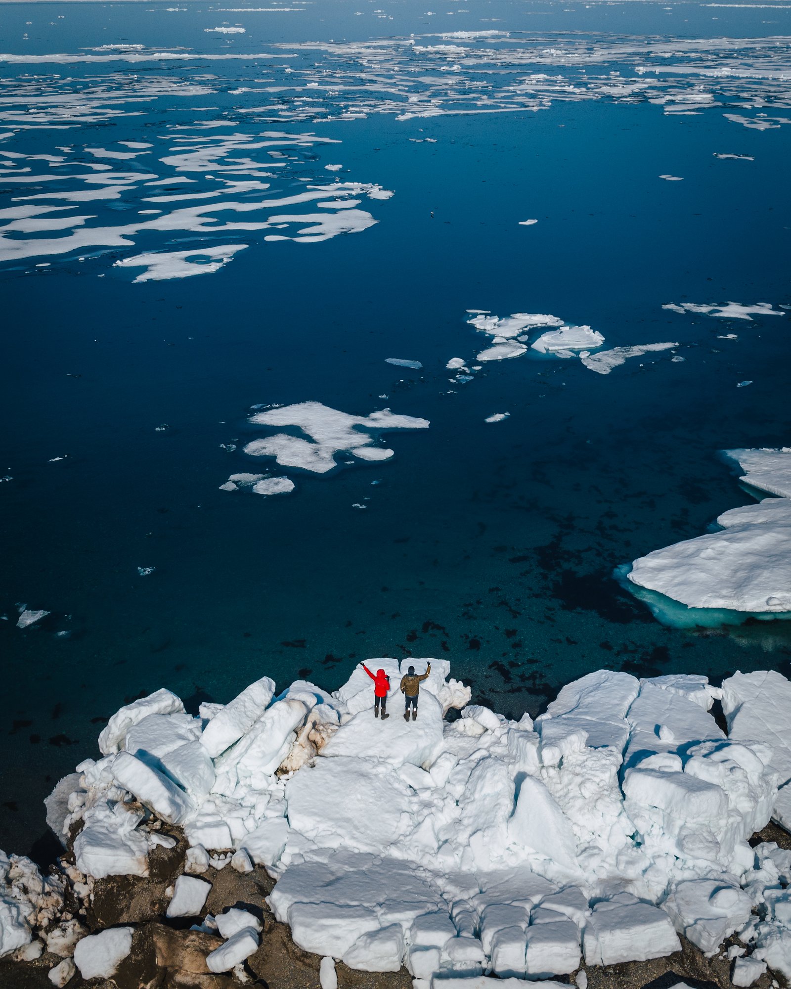 Aerial view of two people standing on a jumble of stranded ice blocks at the shore, floes on dark water
