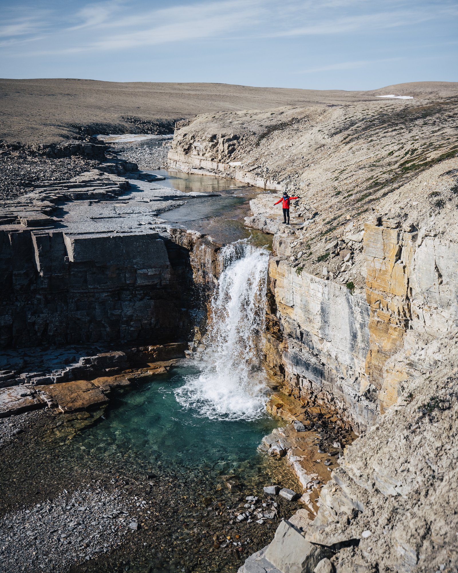The canyon from above: a waterfall and turquoise pool, a hiker in red on the rim