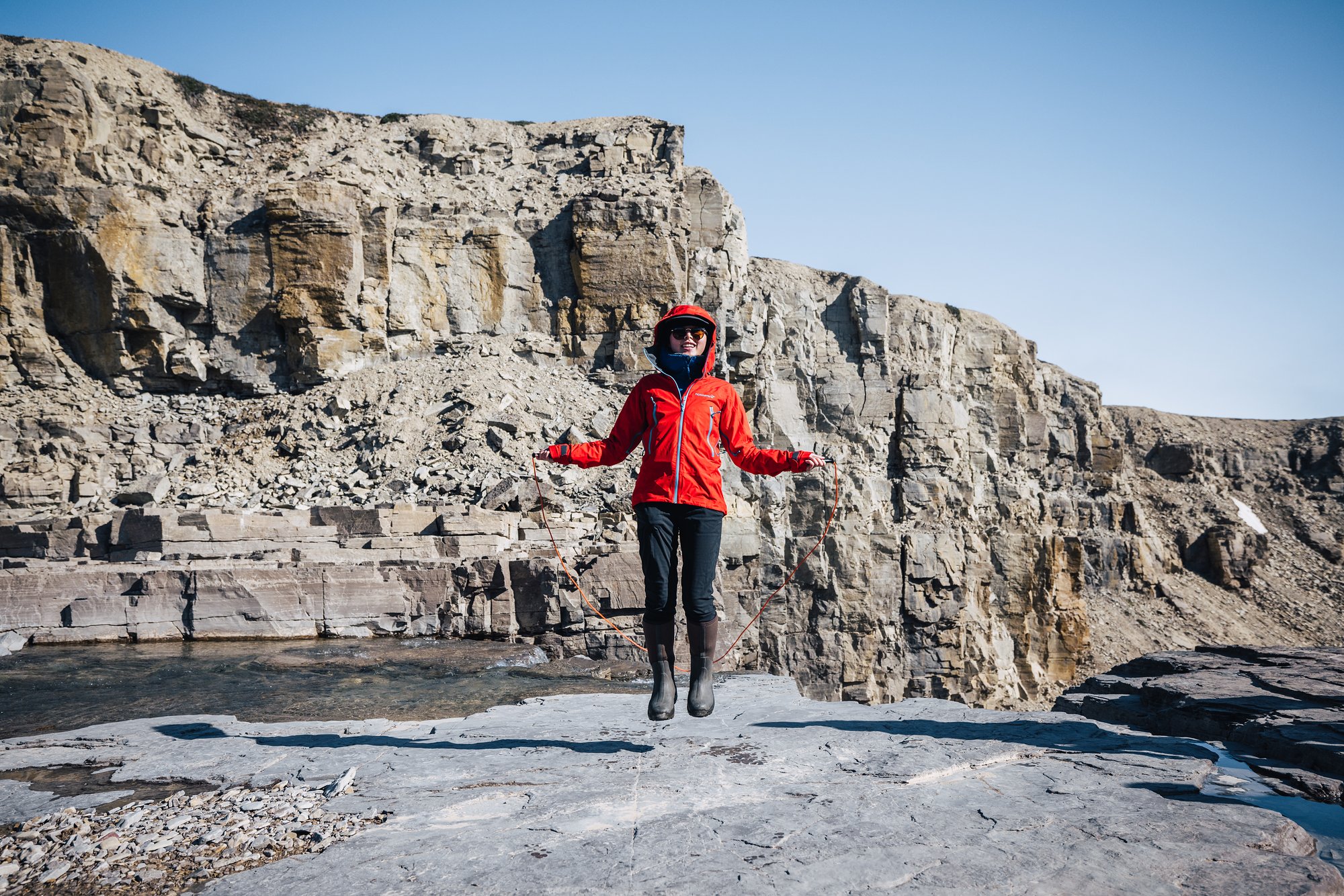 Grace in a red jacket jumping on a rock ledge in front of layered cliffs