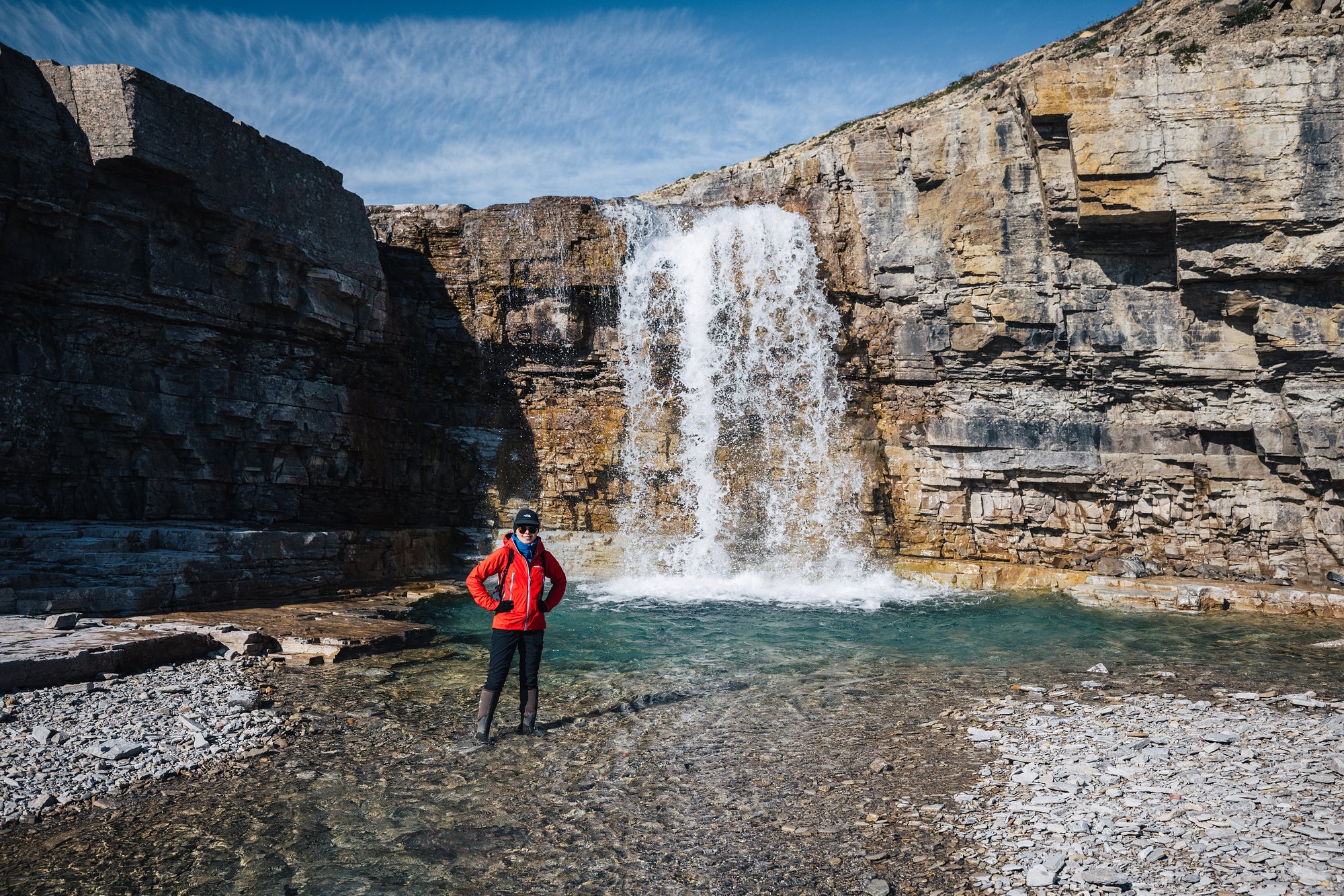 Grace in a red jacket at the foot of a waterfall pouring into a turquoise pool in a layered limestone canyon