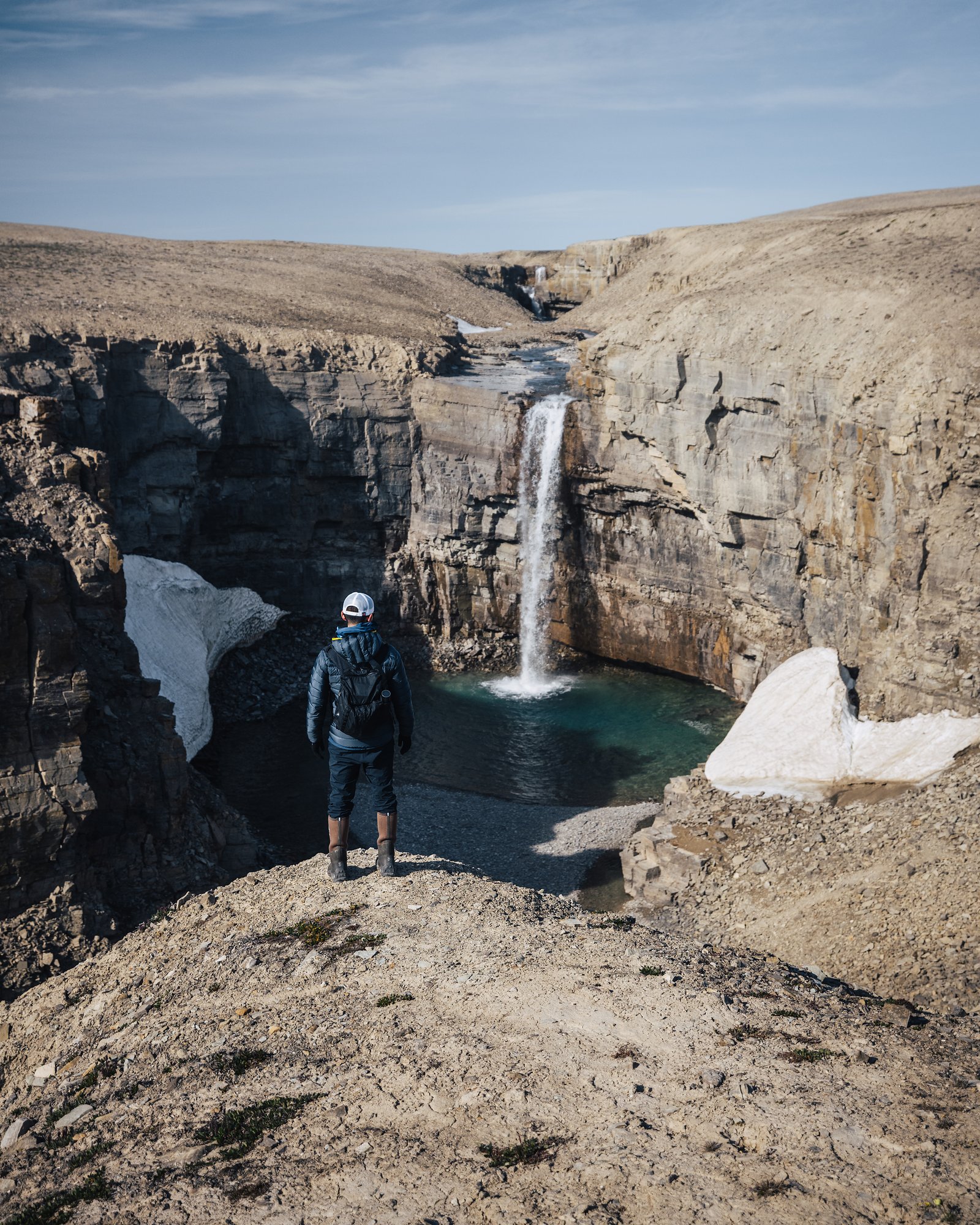 A hiker on the canyon edge looking at a slender waterfall, late snow in the gully