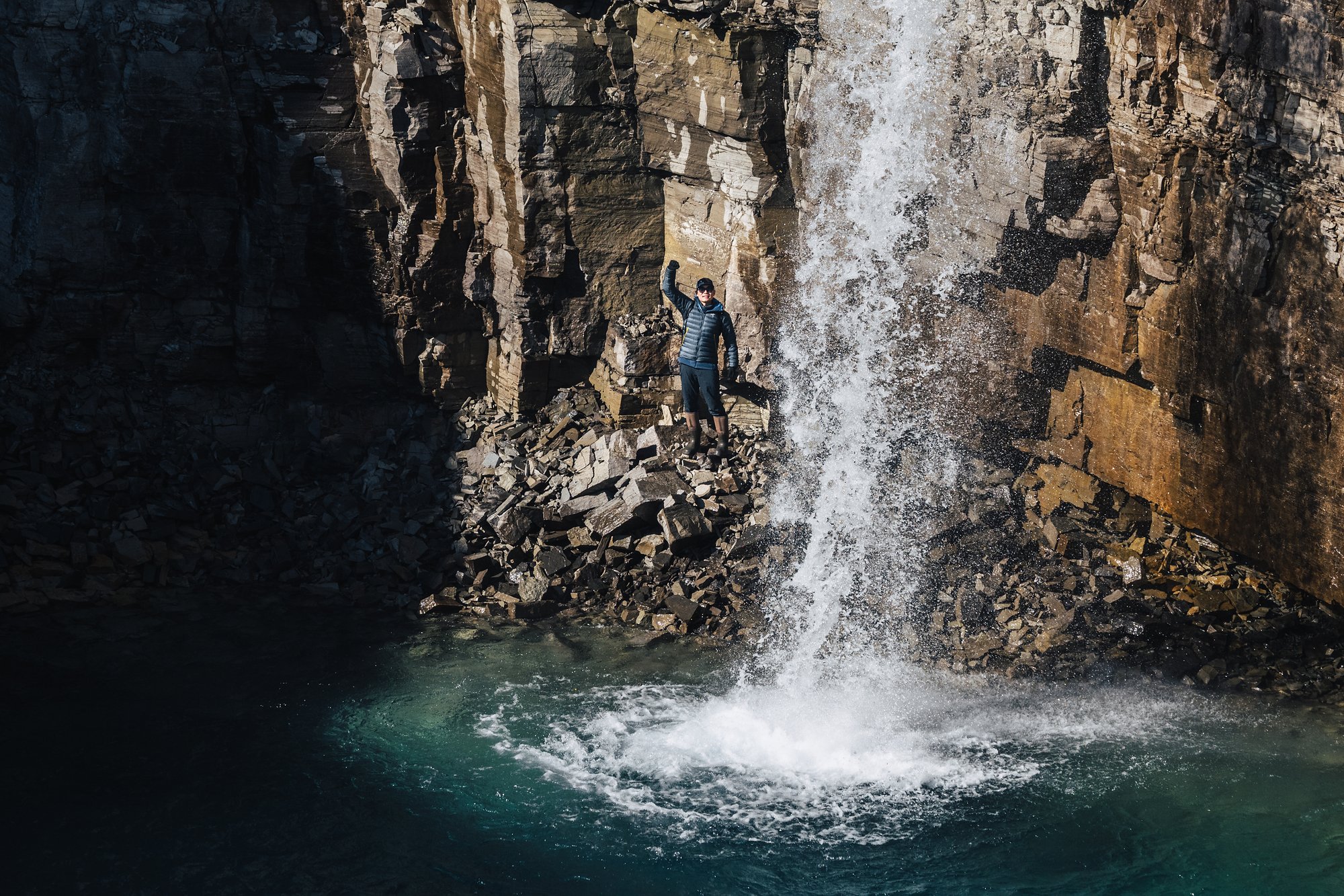A hiker with arms raised on a ledge beside a waterfall in the canyon