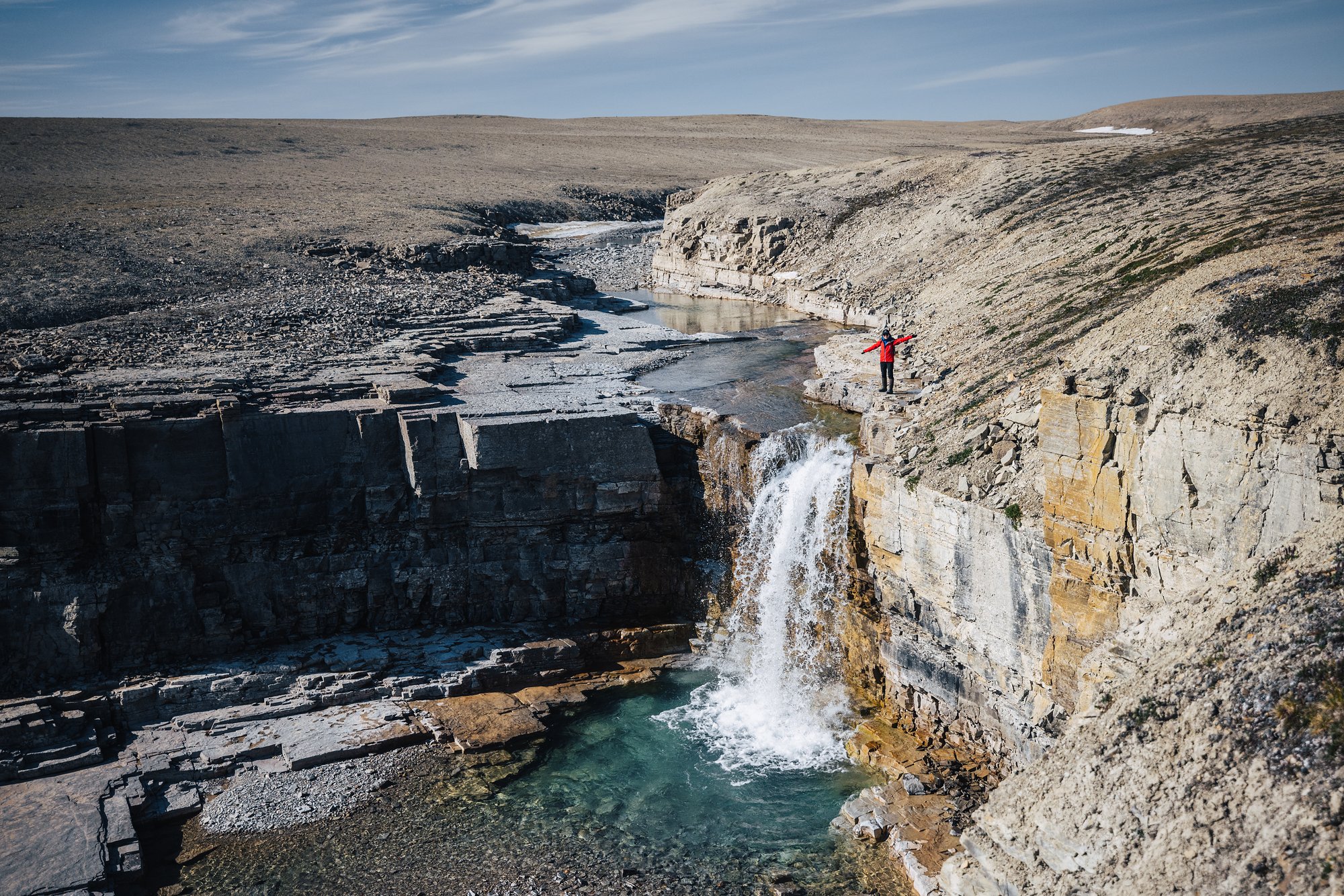 Looking down from the canyon rim at a waterfall and turquoise pool, a hiker in red on the ledge