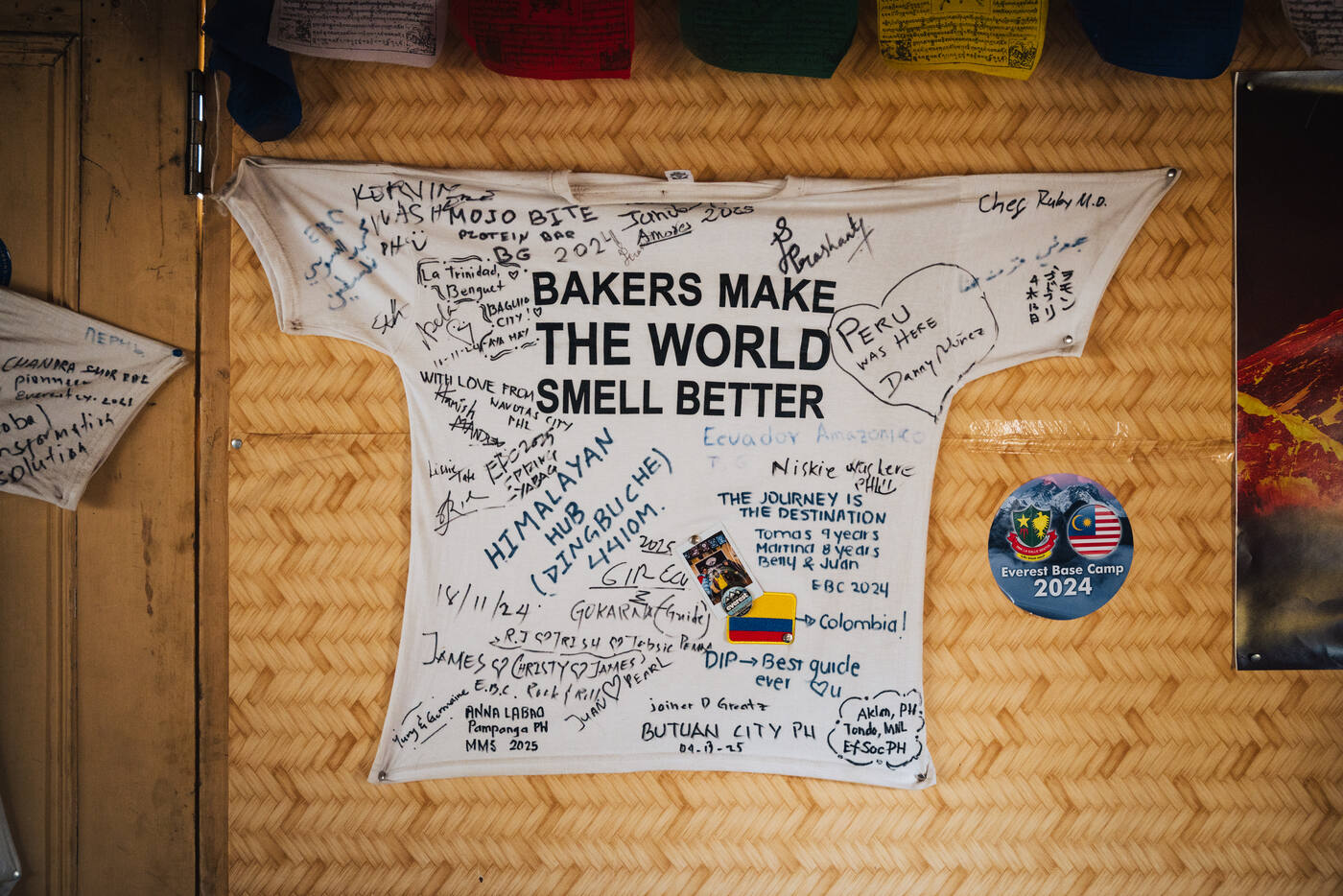 A signed t-shirt reading 'Bakers Make the World Smell Better' pinned to the bakery wall, covered in trekker signatures and messages from around the world