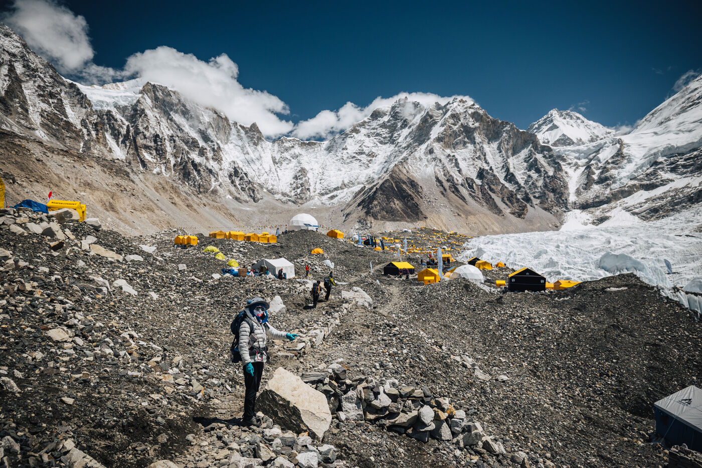 A trekker descending the rocky moraine toward yellow expedition tents at Everest Base Camp, with the snow ridges of Pumori and Nuptse rising behind