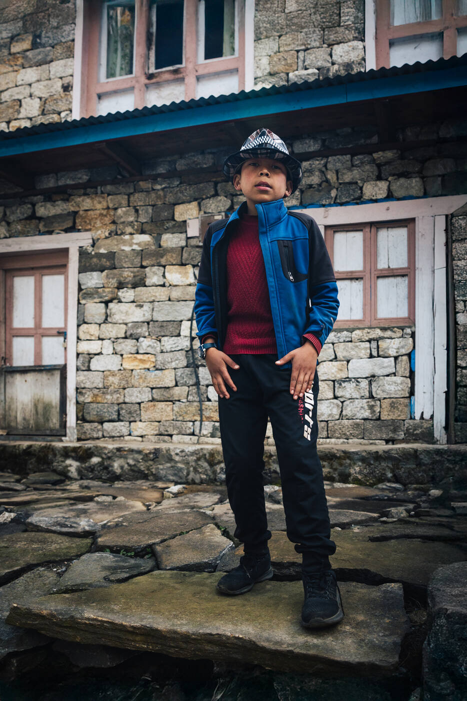 Portrait of a young Namche boy in a blue jacket and checkered bucket hat standing in front of a stone lodge