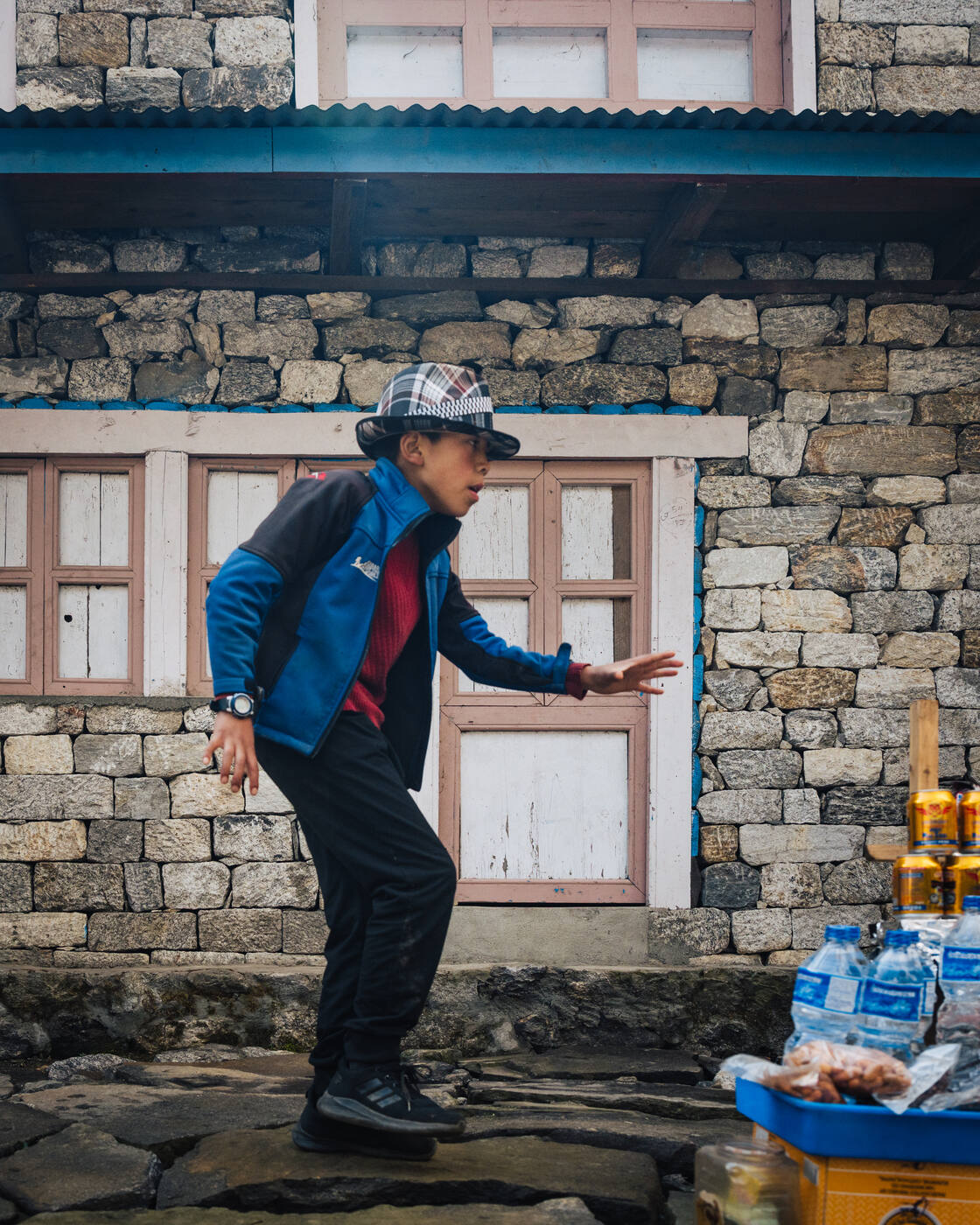 A young Namche boy in a blue jacket and bucket hat reaching toward a small streetside snack stand