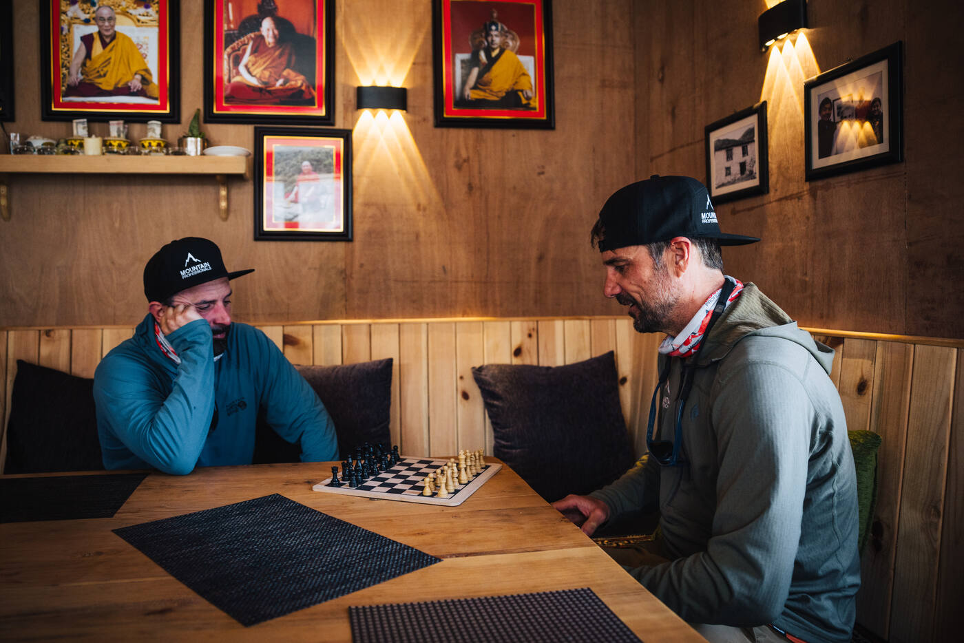 Two trekkers playing chess at a warm-lit lodge table — Buddhist portraits on the wall, wooden paneling behind