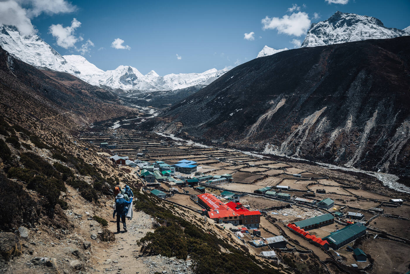 Trekker descending the Nangkartshang ridge above Dingboche — colorful red-roofed lodges on the valley floor below, snow peaks across the horizon
