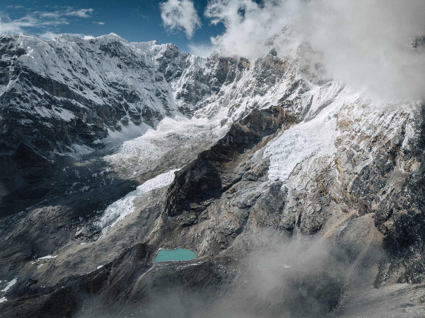 Drone view of a turquoise glacial meltwater pool at the foot of a snow-streaked rock face, clouds curling around the peak above