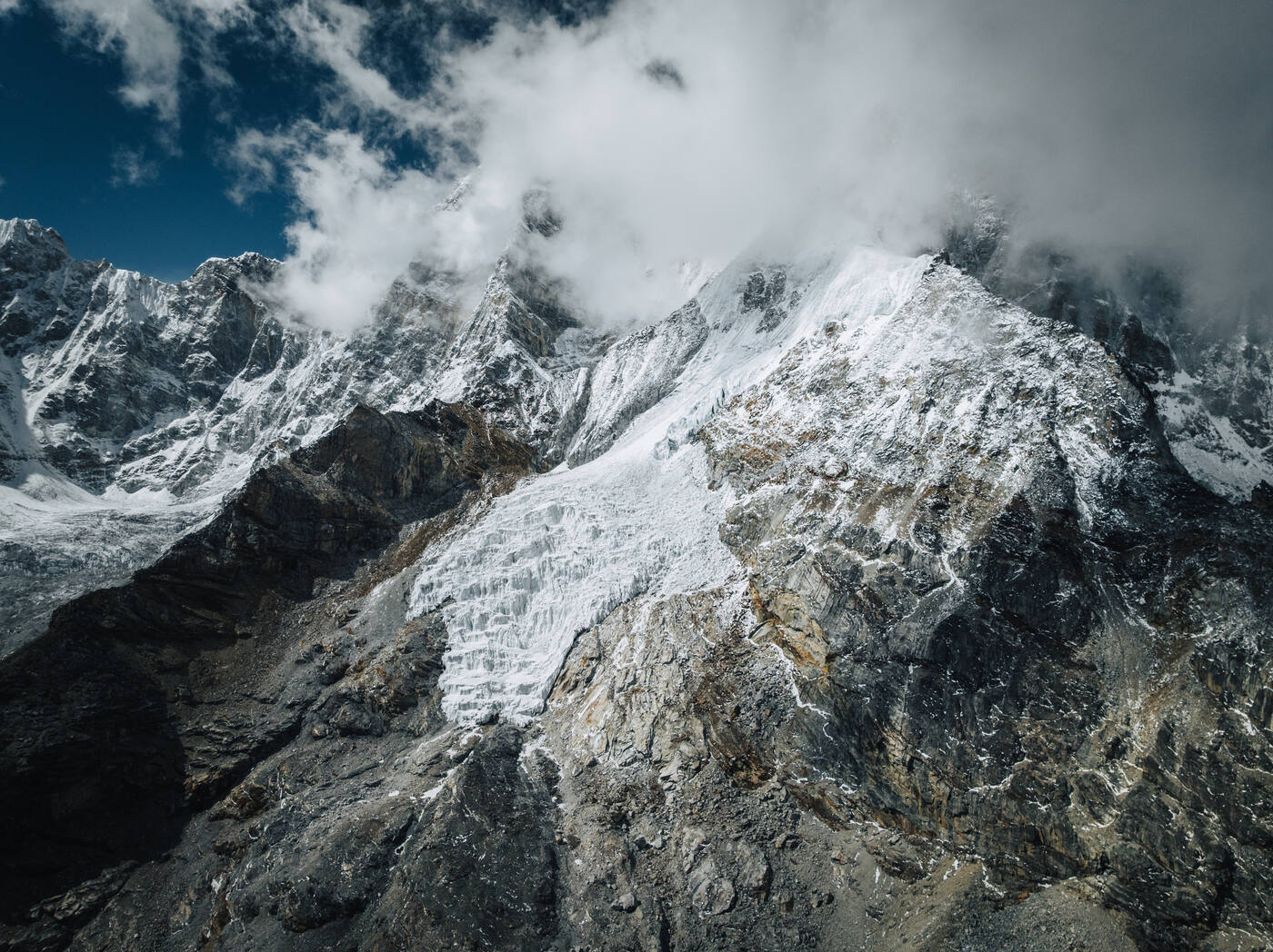 Drone close-up of a snow-clad rock face on Pumori — clouds spilling over the ridge with blue sky above