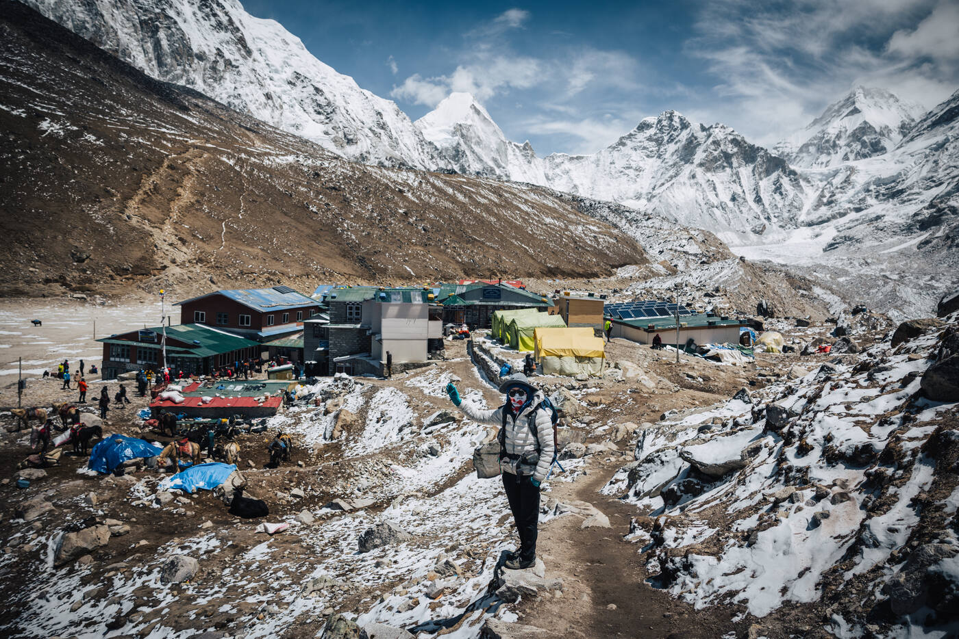 A trekker posing in front of Gorak Shep's tin-roof lodges, snow-streaked peaks rising behind on the high glacial flat
