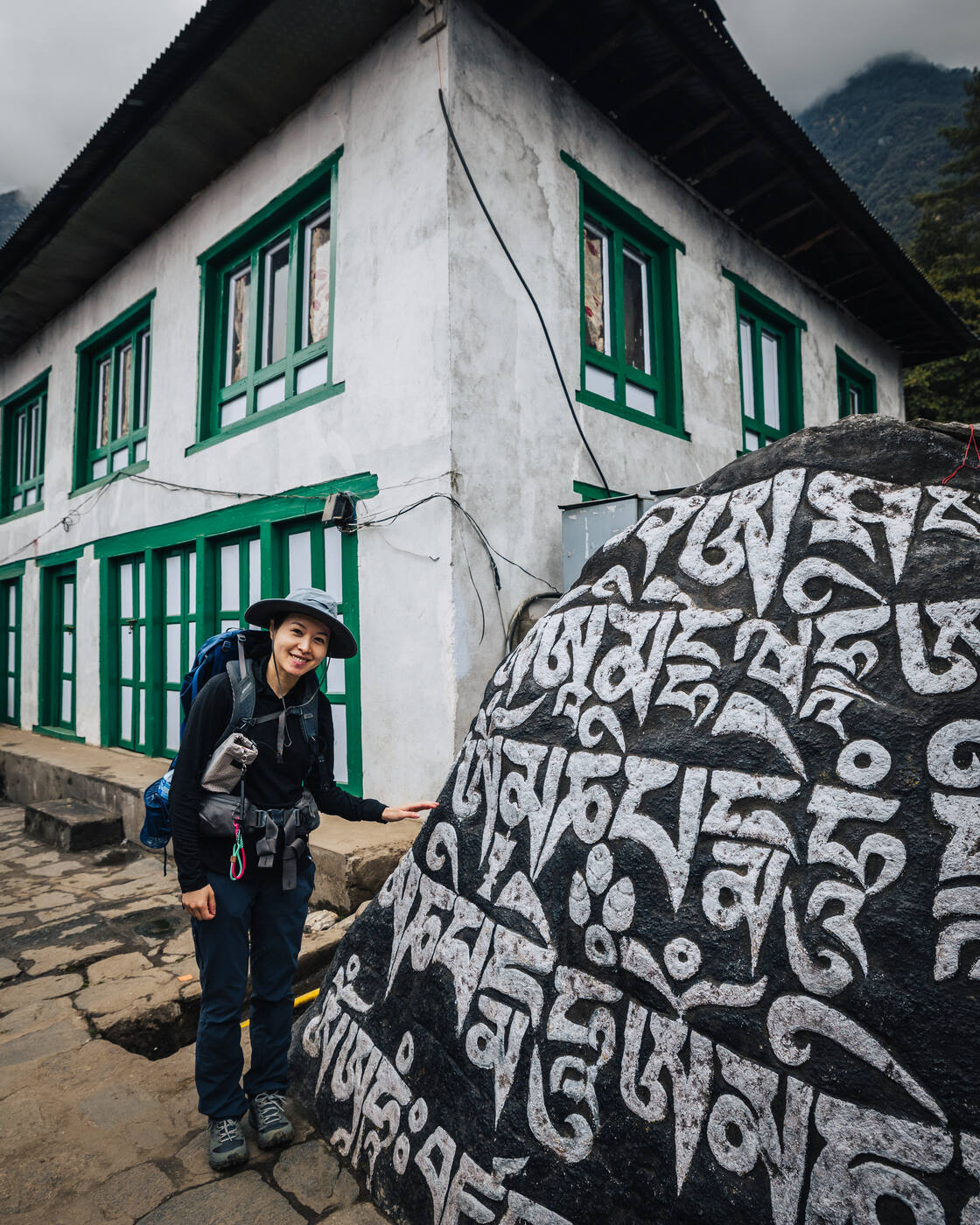 Grace standing next to a large carved mani stone with Tibetan script beside a white building
