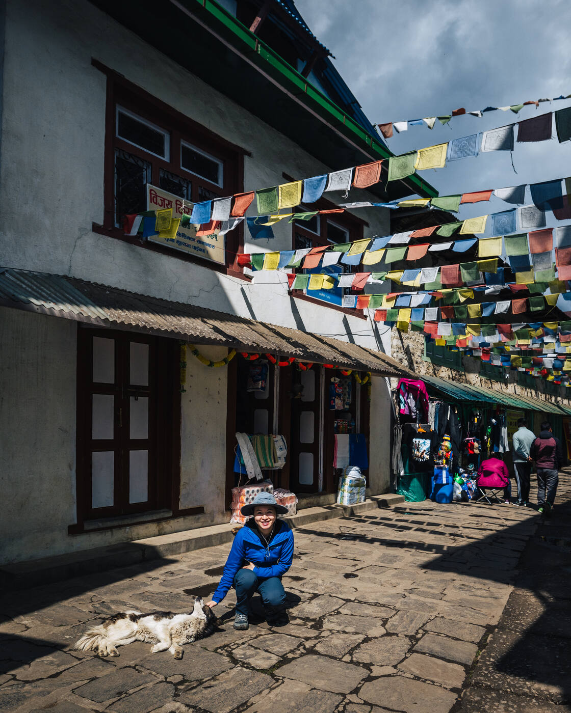 Grace crouched down petting a dog on a Lukla street with colorful prayer flags strung above the shops