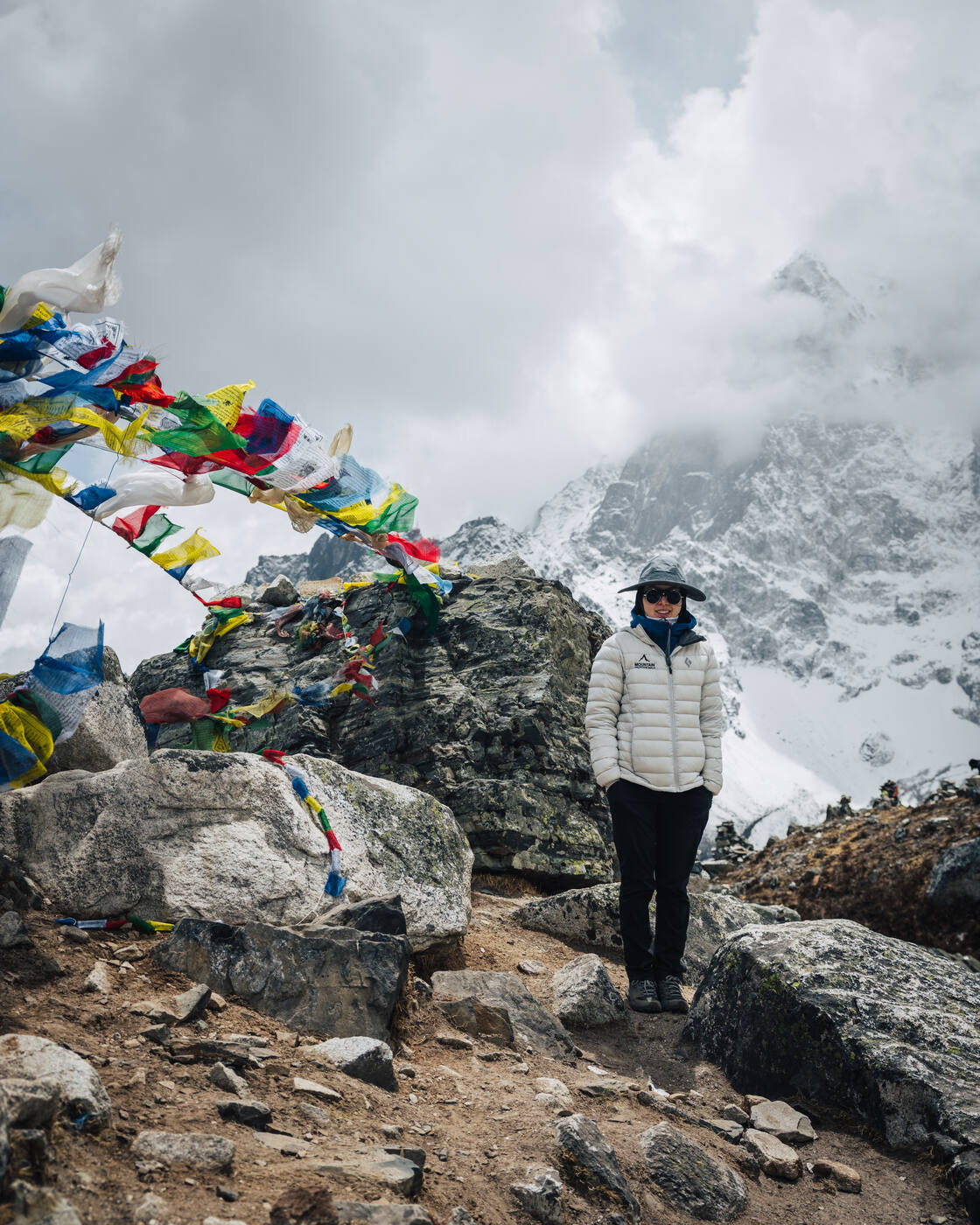 Grace in a white puffy beside chaotic prayer flags on the Thukla Pass, cloud-wrapped snow peaks behind