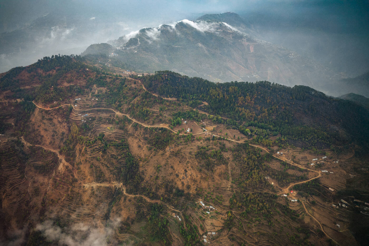 Aerial view of Himalayan foothills with terraced slopes and snow-capped peaks in the distance
