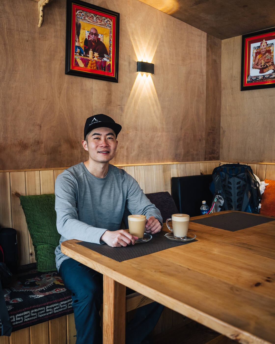 Jason seated at the Dingboche Inn with two lattes on a wooden table, Tibetan portraits and a wall light behind, backpack on the bench beside him