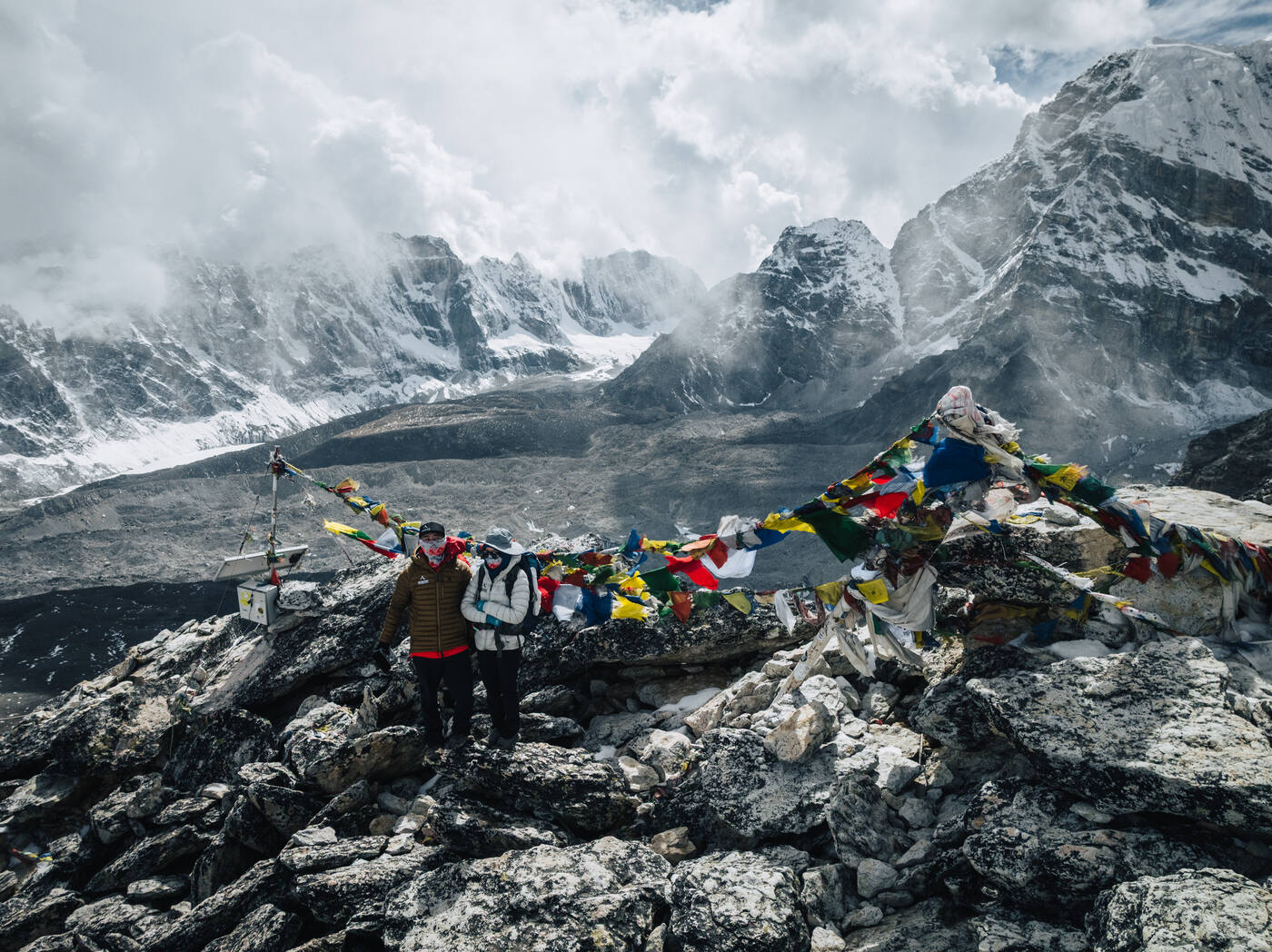 Jason and Grace standing on a Kala Patthar summit boulder draped in chaotic prayer flags, cloud-wrapped peaks behind