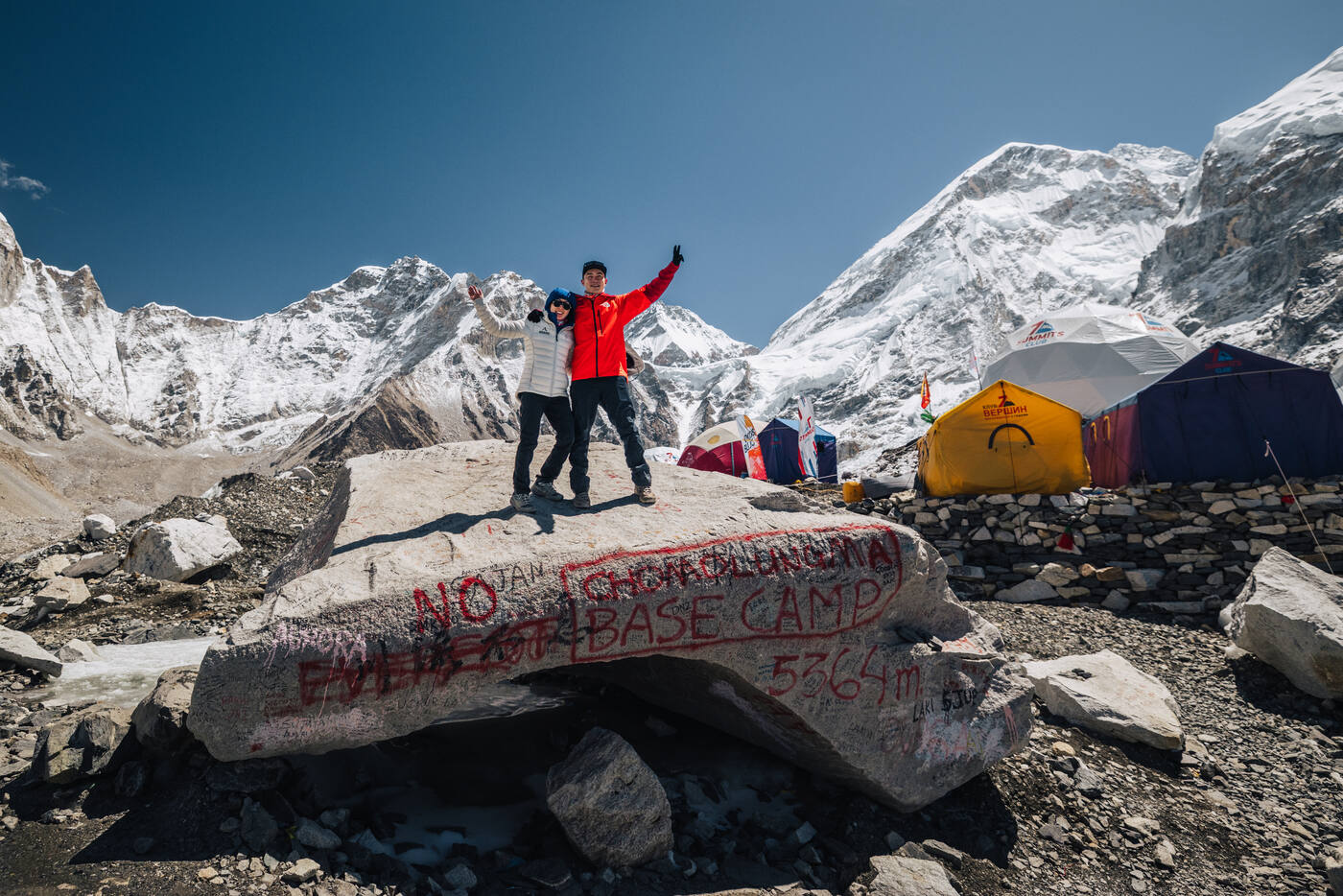 Jason and Grace triumphant on the spray-painted 'EVEREST BASE CAMP 5364m' Trekker's Rock, expedition tents and the Khumbu Icefall behind