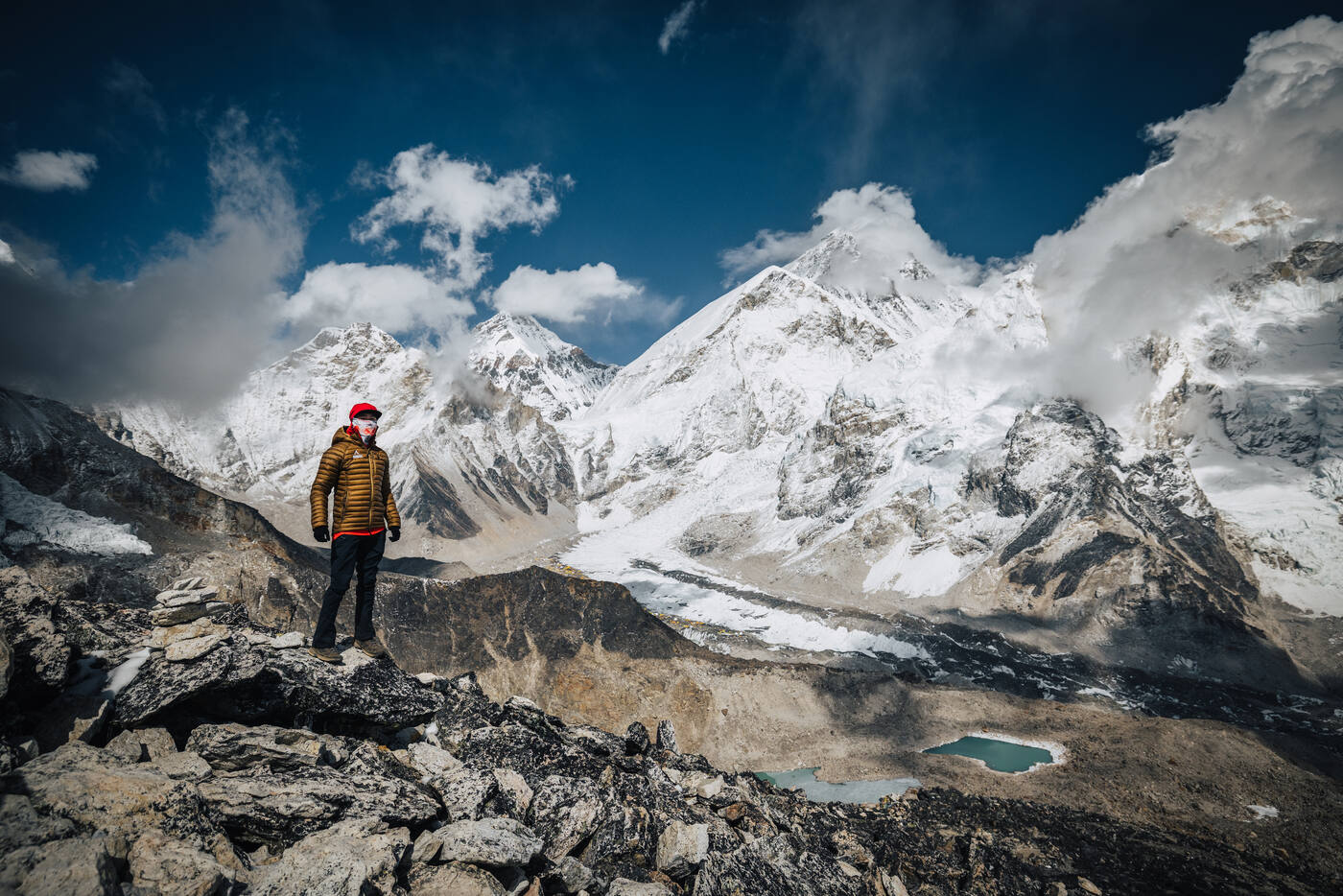 Jason in a yellow-striped puffy jacket on a rocky summit, the massive Everest and Nuptse wall filling the sky behind with a turquoise meltwater pool below