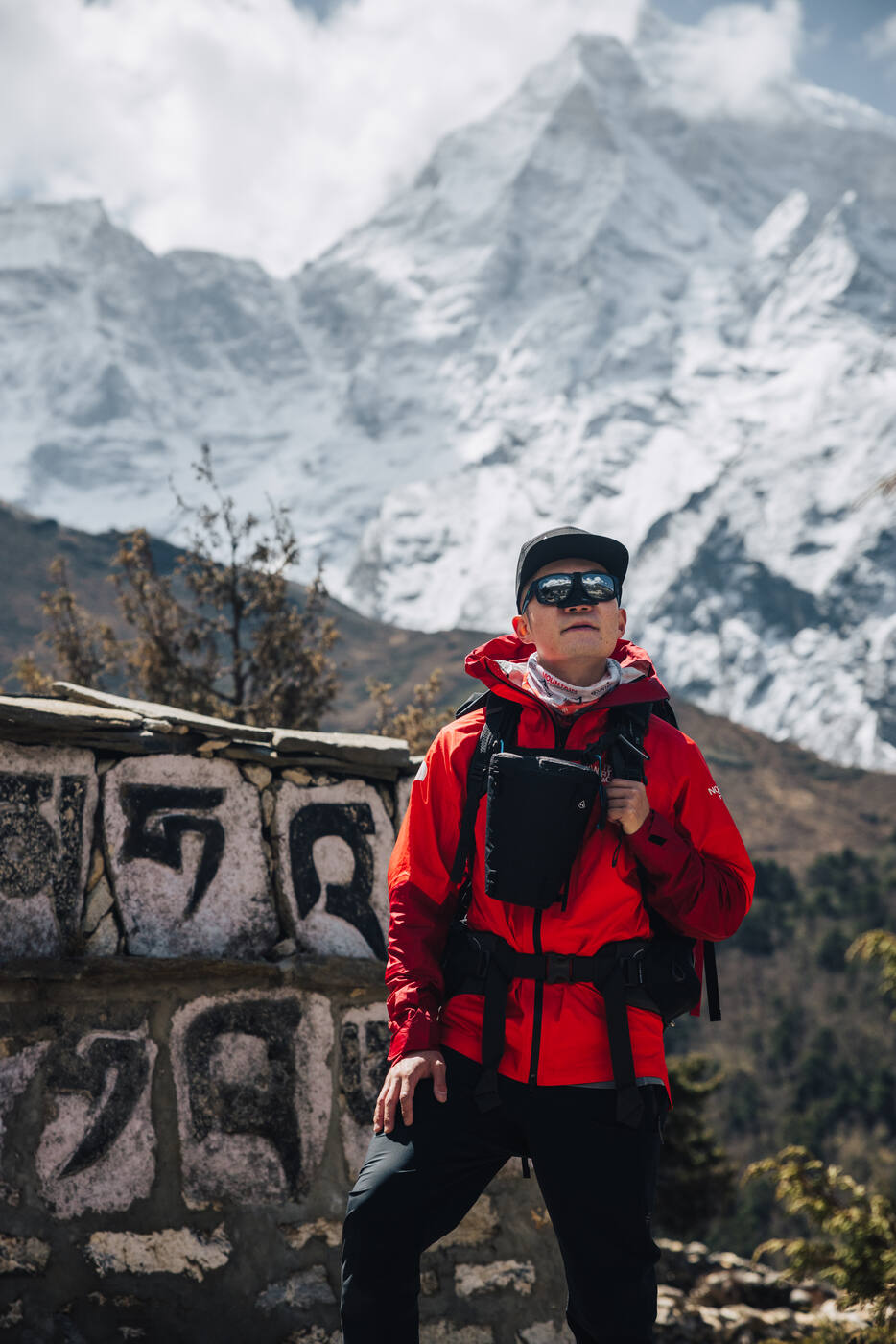 Jason in a red North Face jacket and sunglasses standing beside a painted mani rock, snow ridges behind