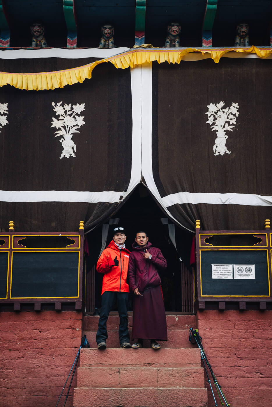 Jason in a red jacket on the monastery steps with a young monk in maroon robes outside the Tengboche gateway