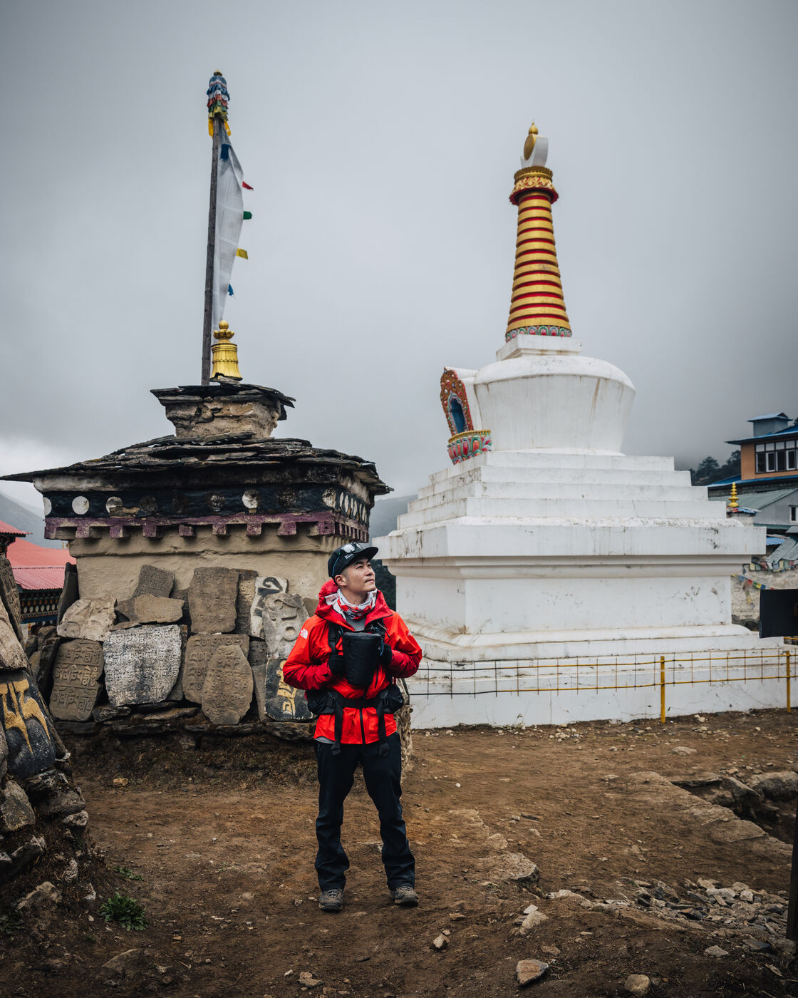 Jason in a red puffy jacket standing before the Tenzing Norgay Stupa — a small mani shrine on the left and a tall white-and-gold stupa on the right under an overcast sky