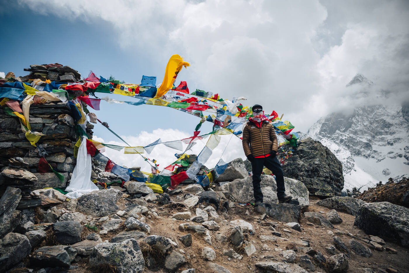 Jason in a yellow-brown puffy at the Thukla Pass memorial, layered prayer flags streaming across a rocky cairn field with cloud-wrapped peaks behind