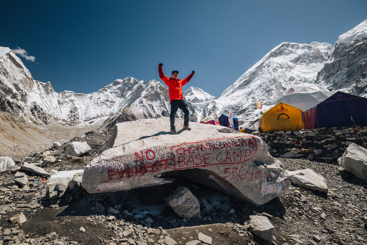 Jason alone on the spray-painted Trekker's Rock boulder with arms raised in triumph, the 'EVEREST BASE CAMP 5364m' sign visible below, expedition tents and snow peaks behind
