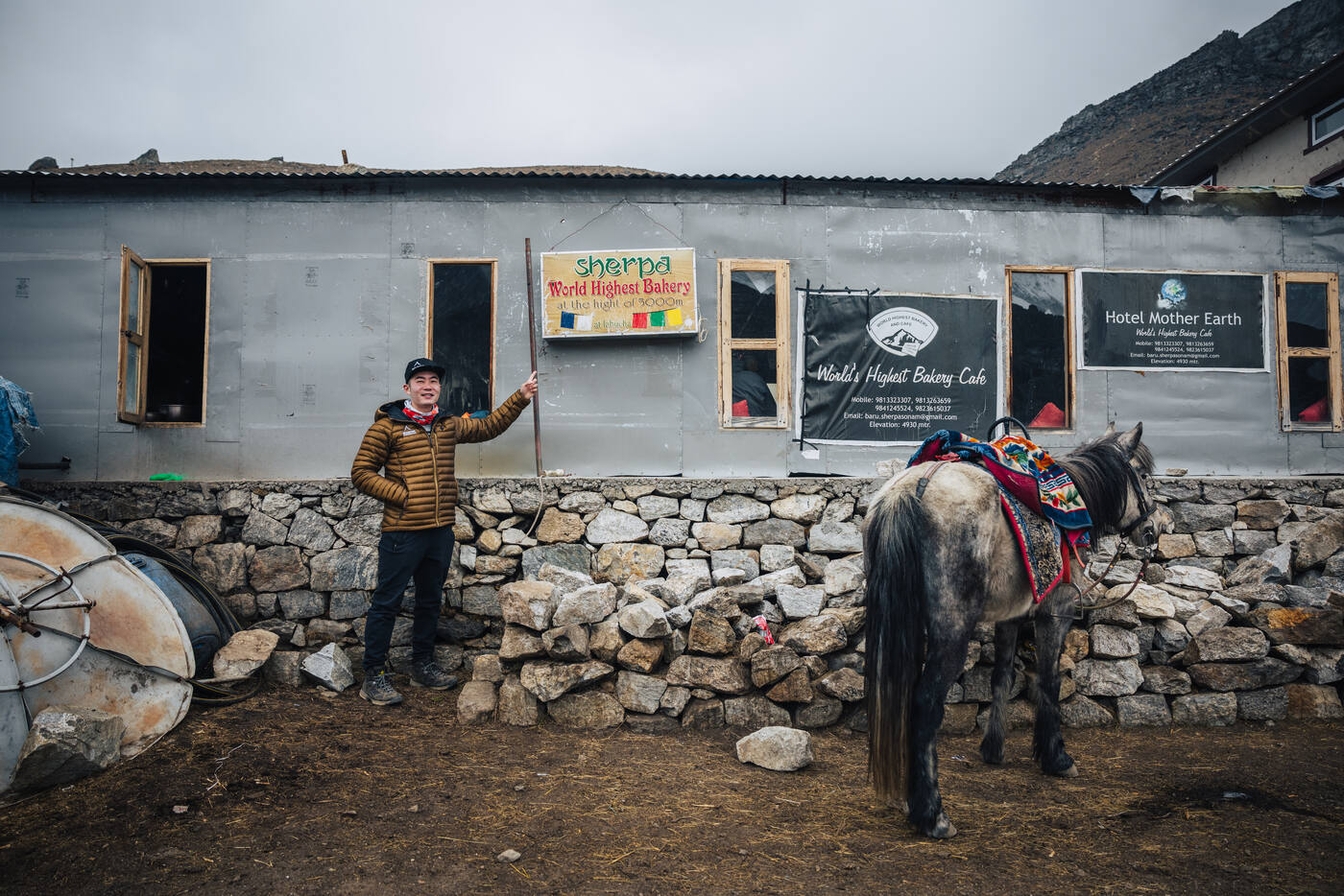 Jason posing beside the Sherpa World's Highest Bakery and Hotel Mother Earth signs at Lobuche, a tethered horse beside the stone wall
