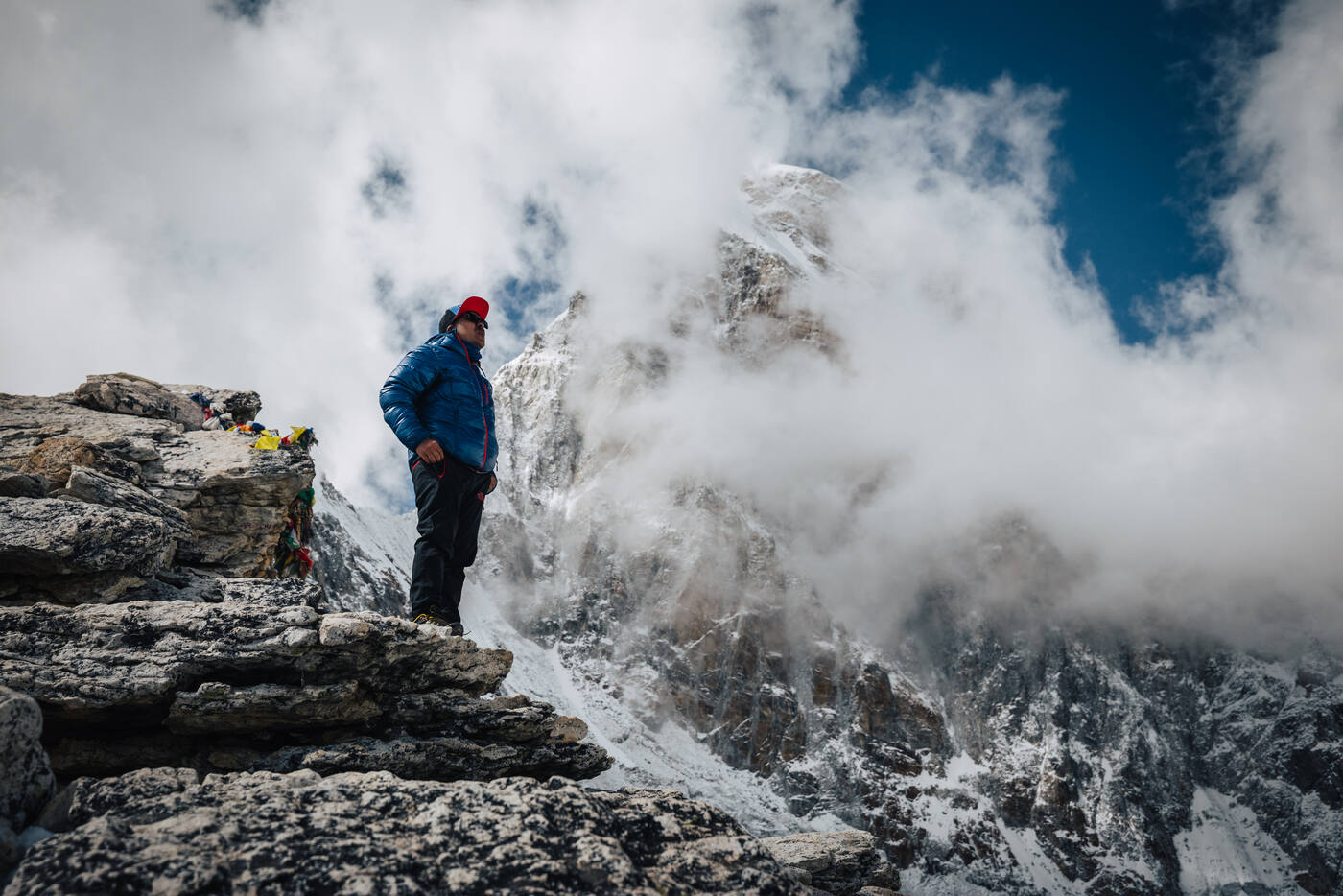 A trekker in a blue jacket and red cap on the rocky Kala Patthar summit, clouds billowing around the snow-pyramid peaks behind