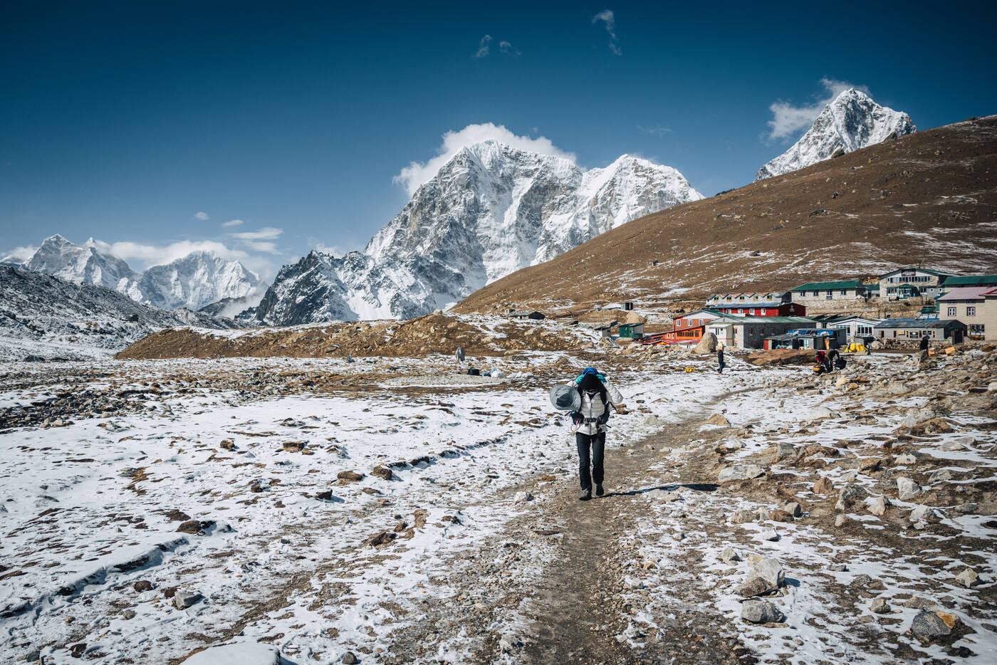 Trekker with a backpack walking a snow-dusted moraine toward a cluster of tin-roof lodges, Cholatse's dark pyramid and other peaks behind — arriving in Lobuche